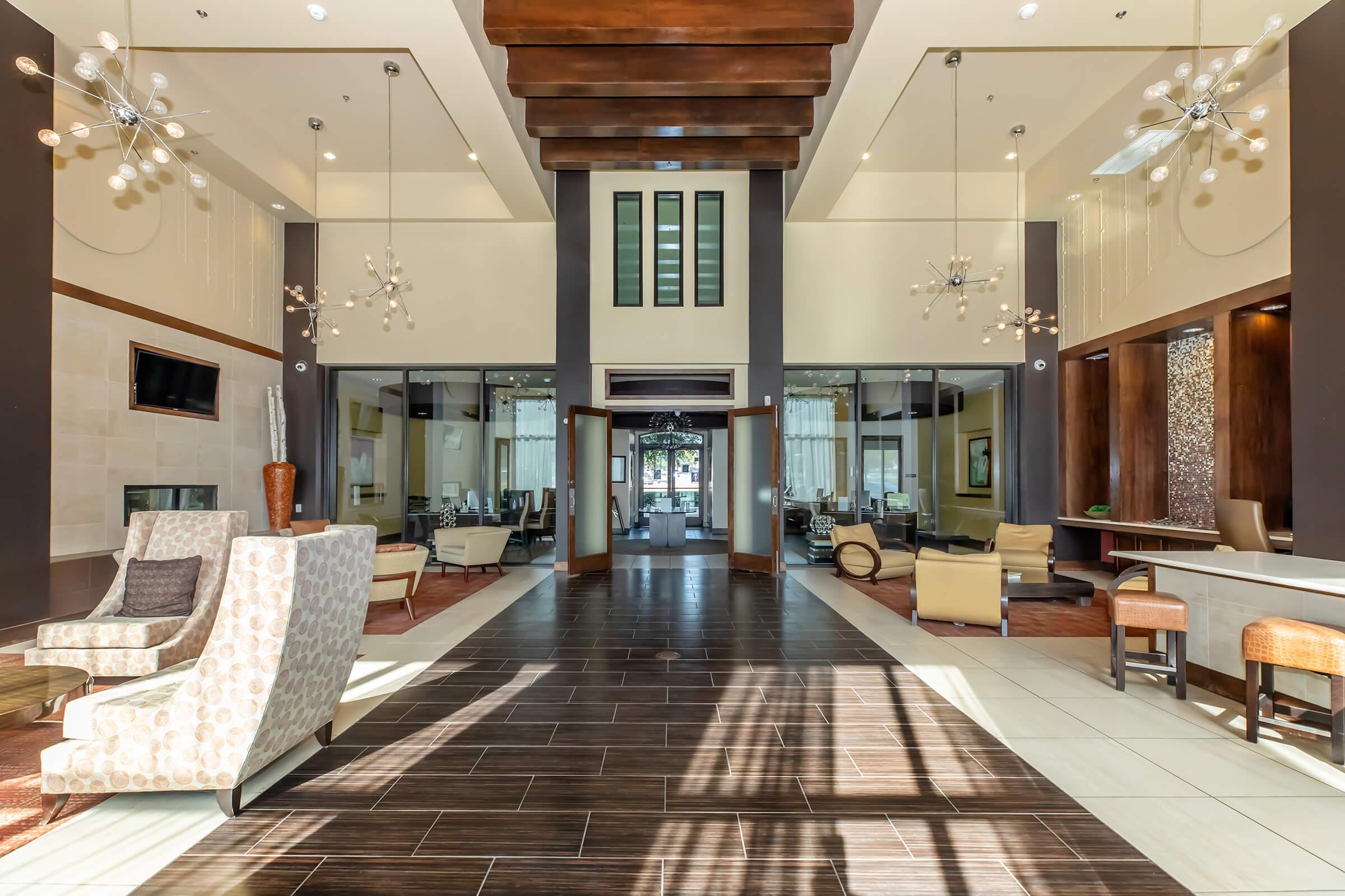 A spacious lobby with modern decor, featuring stylish chairs, a stone fireplace, large windows allowing natural light, and elegant lighting fixtures. The open space has a polished wooden ceiling and a welcoming atmosphere, with reception areas visible in the background.
