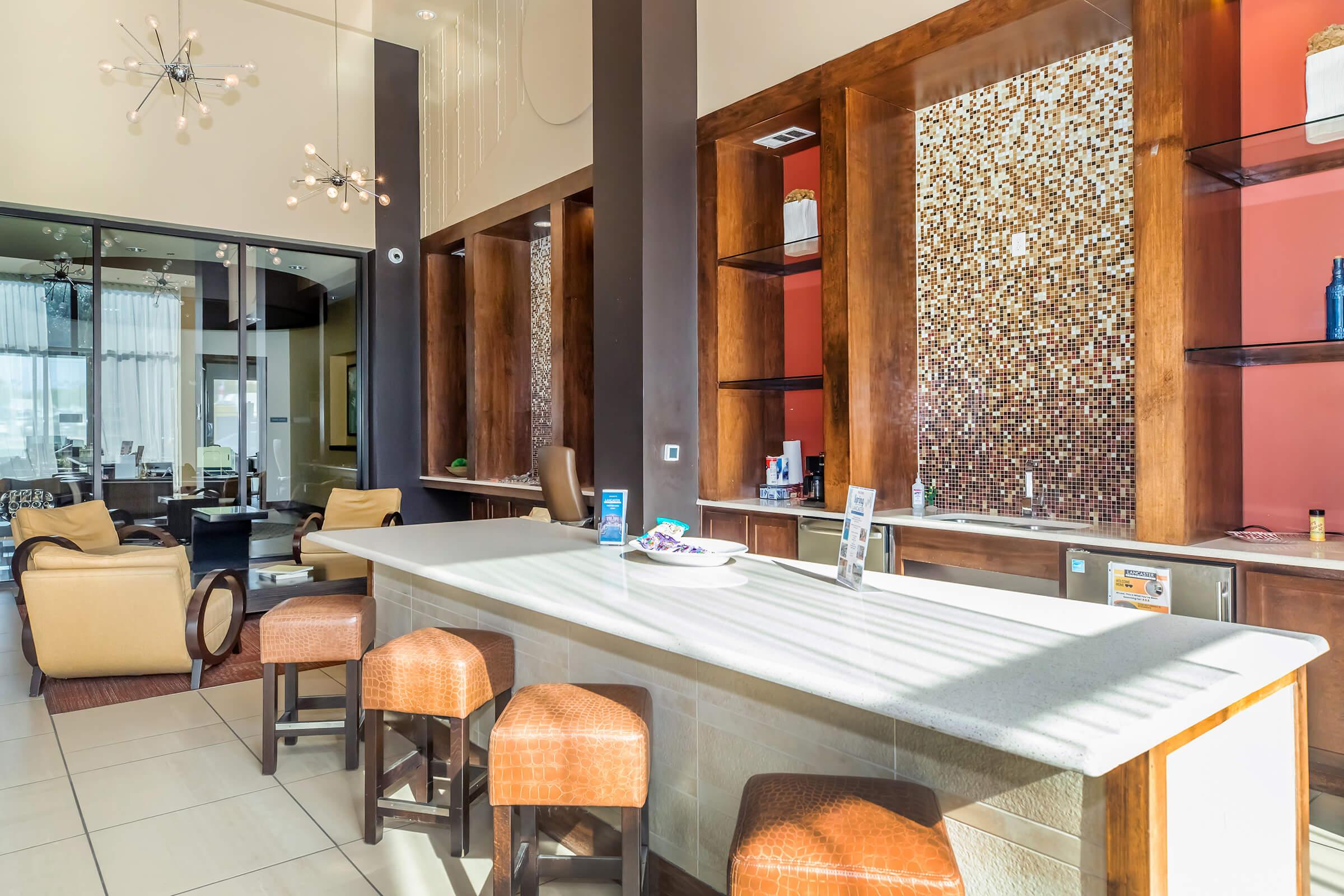 Modern lounge area featuring a sleek countertop with bar stools, elegant pendant lighting, and a stylish backsplash. The space is designed with large windows for natural light, comfortable seating in the background, and decorative accents, creating an inviting atmosphere.
