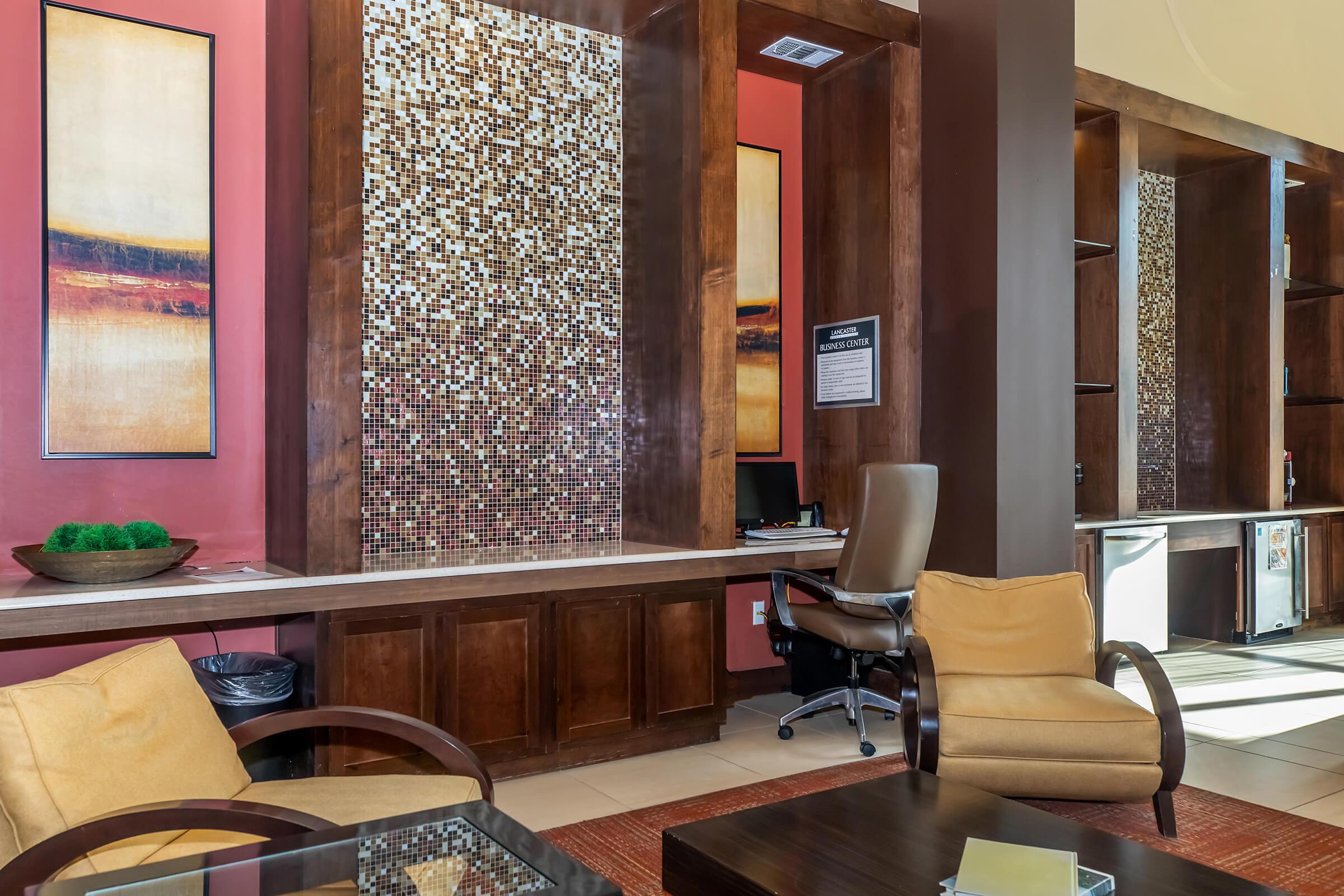 Interior of a modern lobby featuring warm-toned walls, a decorative mosaic feature, and a comfortable seating area with two armchairs. A desk with a chair is positioned against the wall, and shelves are visible. Natural light enhances the inviting atmosphere.