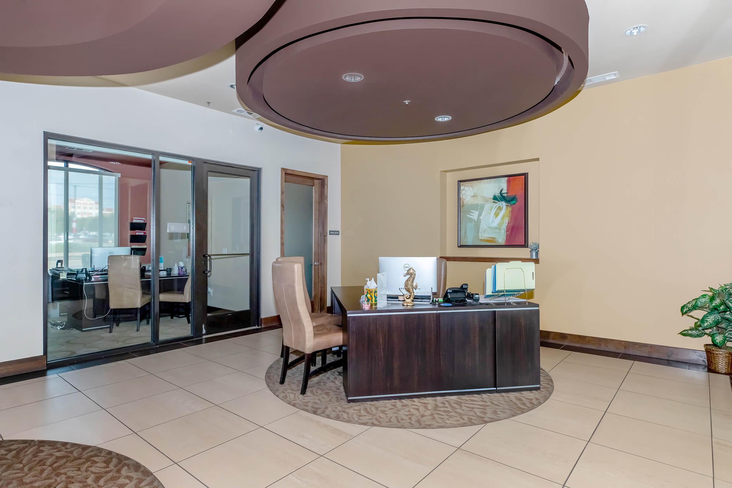 Modern office interior featuring a desk with a chair, decorative items, and a potted plant. A glass door leads to another office space in the background. The ceiling has a unique circular design, and a colorful abstract painting adorns the wall. The flooring is tiled with a carpeted area.