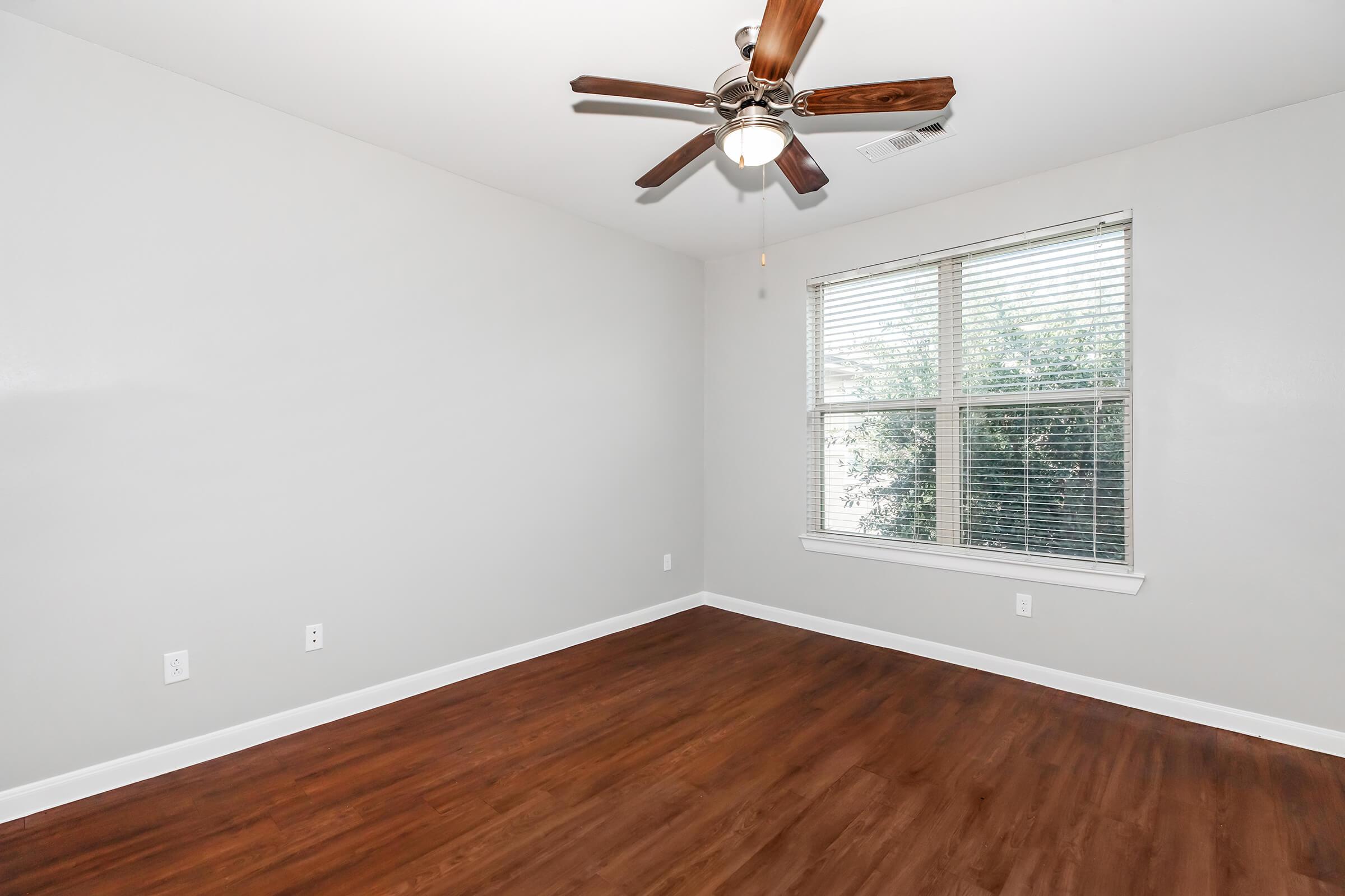 A clean, empty room with a light gray wall and hardwood flooring. The ceiling fan has wooden blades. A window with white blinds lets in natural light, revealing greenery outside. The room appears spacious and ready for furnishing.