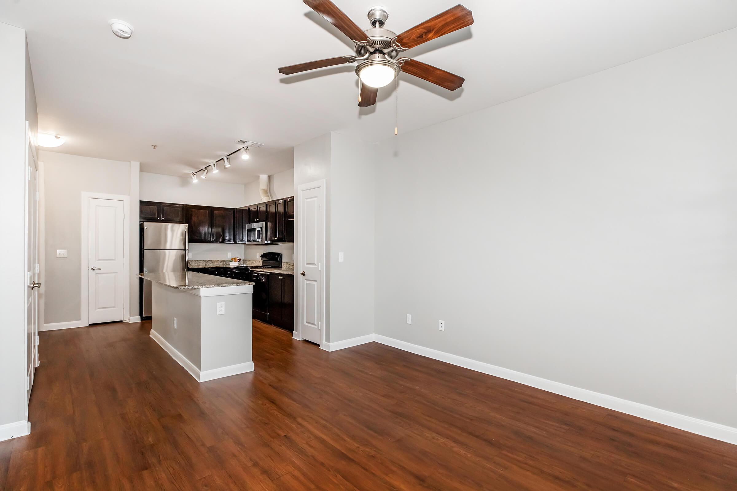 Spacious modern living area featuring hardwood floors, a ceiling fan, and a kitchen with dark cabinetry and stainless steel appliances. Bright natural light illuminates the room, creating an inviting atmosphere. The area is open and versatile, suitable for various furniture arrangements.