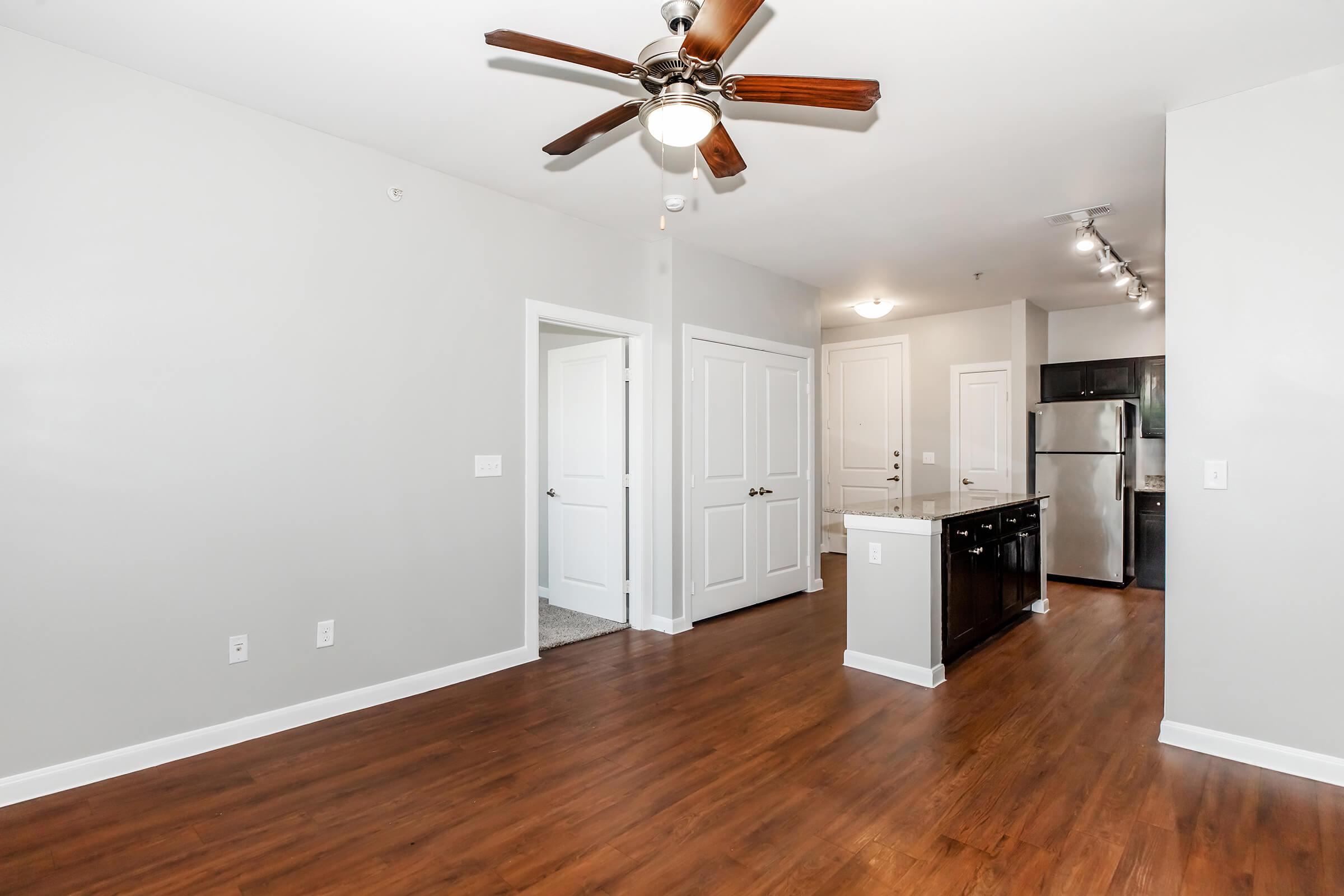 A well-lit, modern apartment interior featuring a living area with hardwood flooring, a ceiling fan, and an open kitchen with stainless steel appliances. There are white walls and doors, and a doorway leading to another room. The overall decor is sleek and contemporary.
