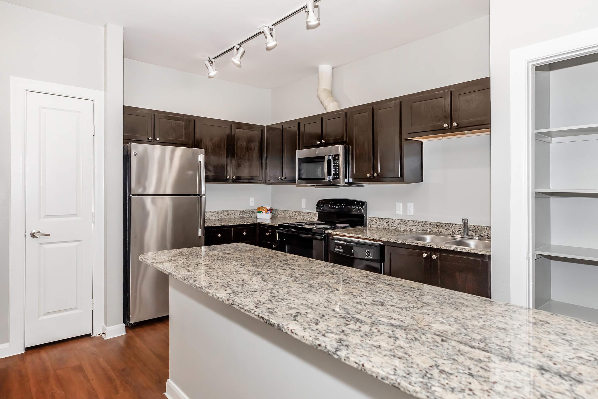 Modern kitchen with dark wood cabinetry and granite countertops. Features stainless steel appliances, including a refrigerator, microwave, and gas stove. An island with bar seating separates the kitchen from the living area, with a pantry visible in the background and natural light from a window.