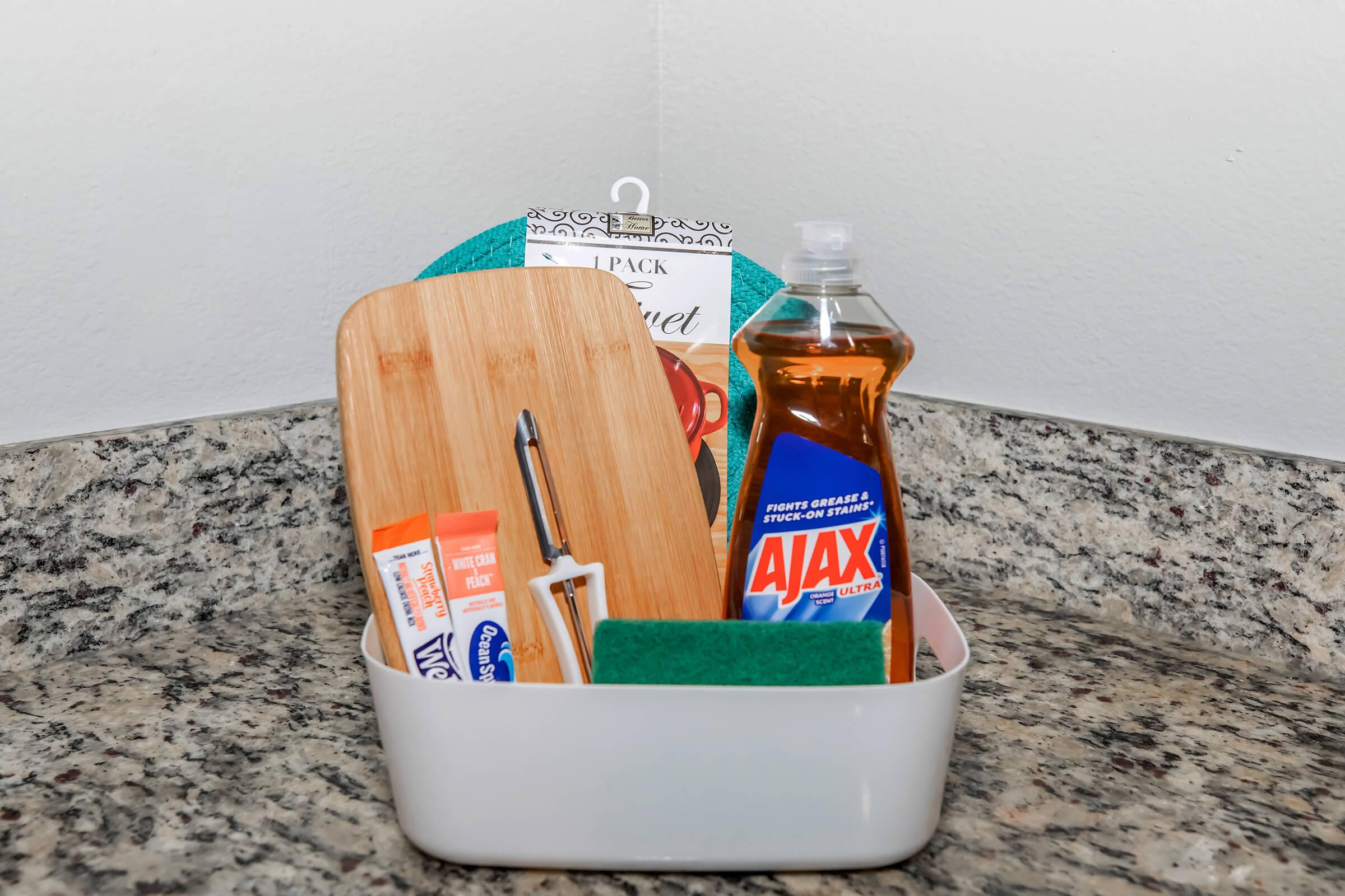 A white container on a granite countertop holding a wooden cutting board, a bottle of Ajax dish soap, a green sponge, a silver pen, and a pack of paper towels.