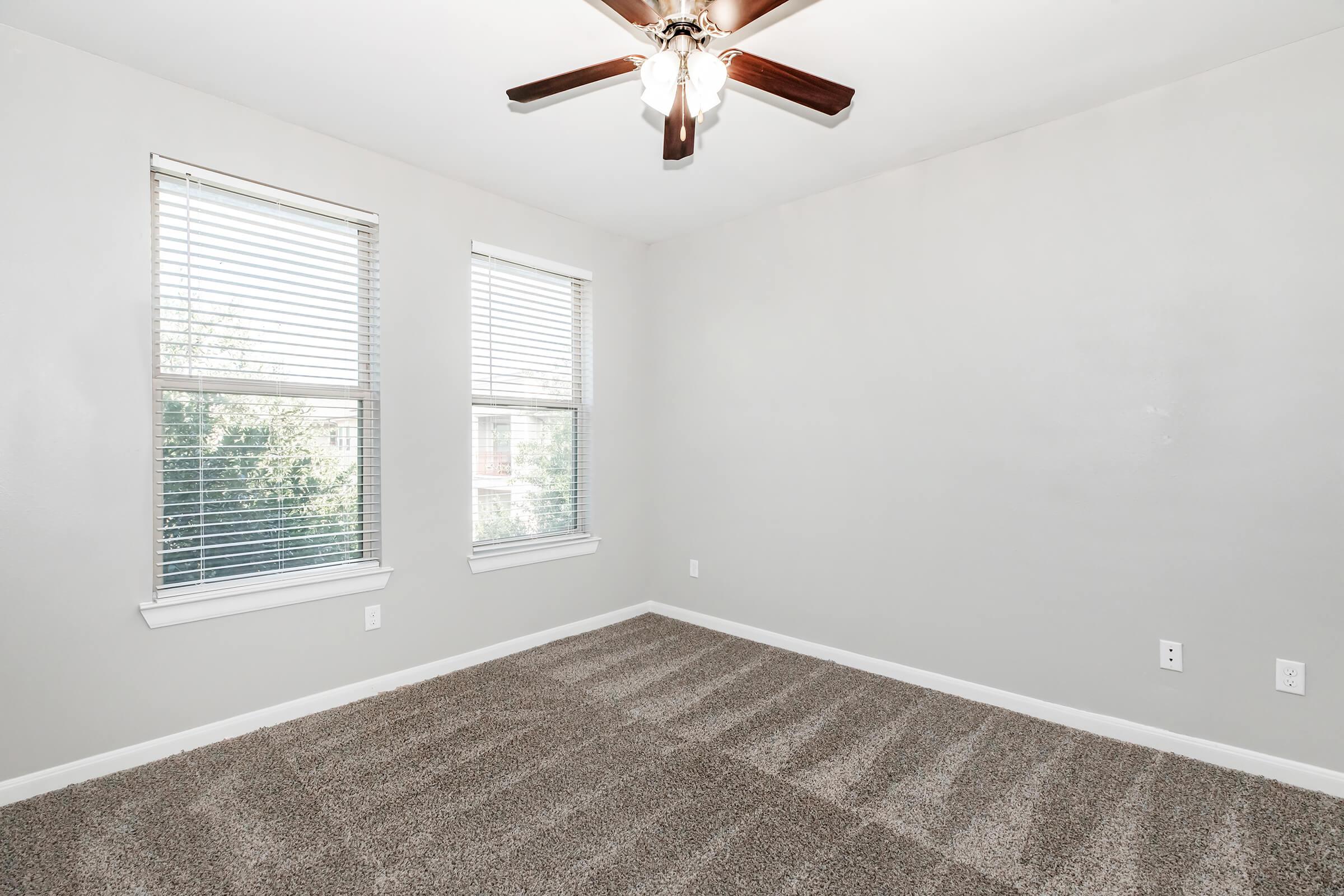 A clean, empty room with light gray walls and a ceiling fan. Two windows with blinds allow natural light to enter, and the floor is covered in plush, tan carpet. The space is bright and uncluttered, ideal for various uses.