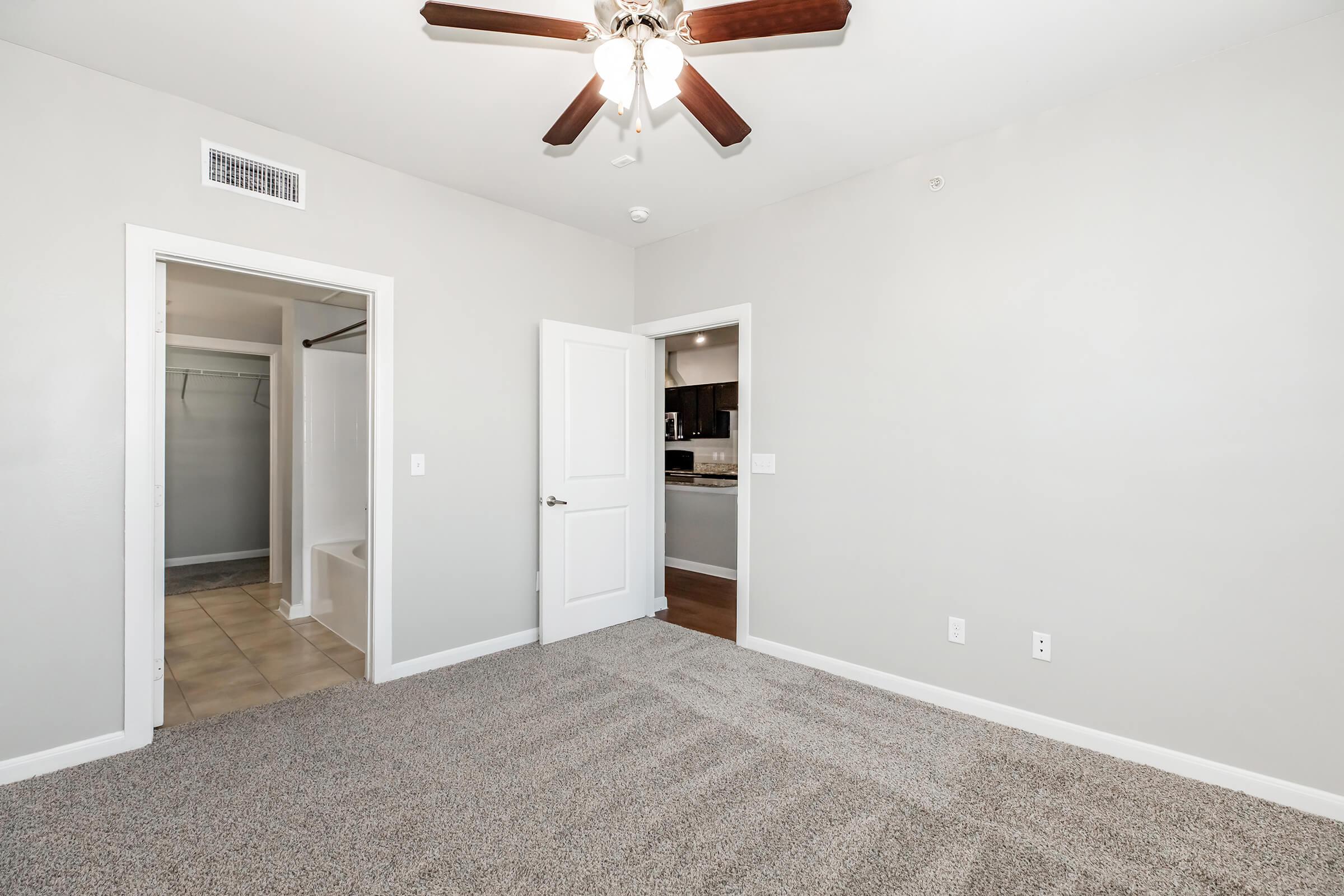 A spacious, well-lit room with light gray walls and a ceiling fan. It features a doorway leading to a closet on the left and an entrance to a kitchen area on the right. The floor is covered with plush carpet, creating a cozy atmosphere.