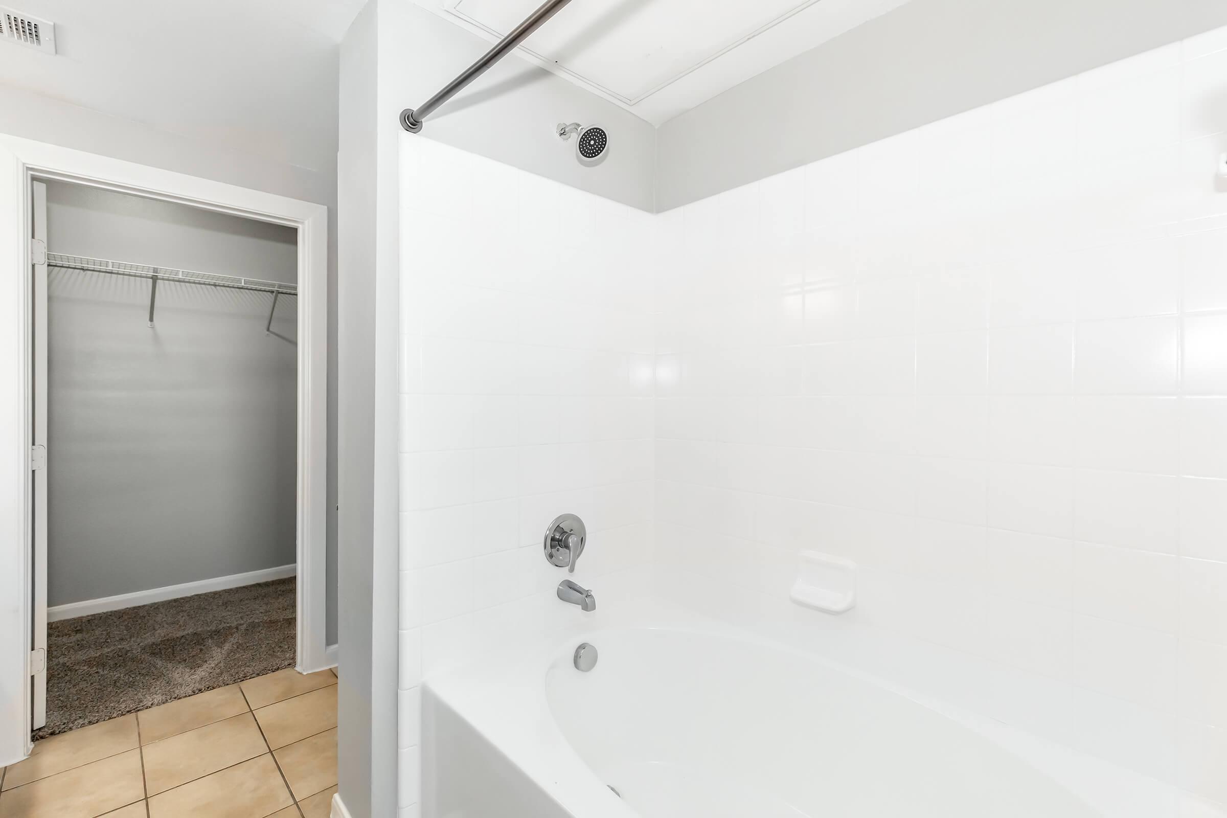 A clean, modern bathroom featuring a white bathtub and shower combination. The walls are painted light gray, and the floor is tiled in a beige tone. In the background, there is a closet with an empty wooden rod and additional space visible, giving an airy and uncluttered feel to the area.