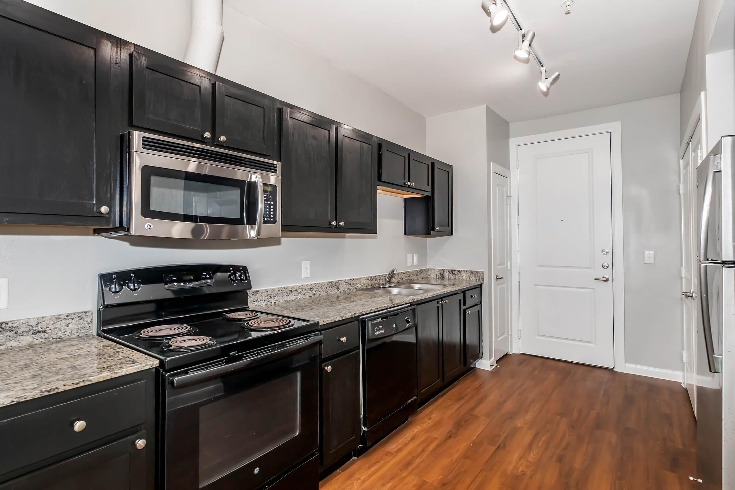 Modern kitchen with dark cabinetry, stainless steel appliances, and granite countertops. Features a range, microwave, and dishwasher. The space has a warm wood floor and neutral-toned walls, with ample lighting from track fixtures. A door leads out, adding to the room's functionality.