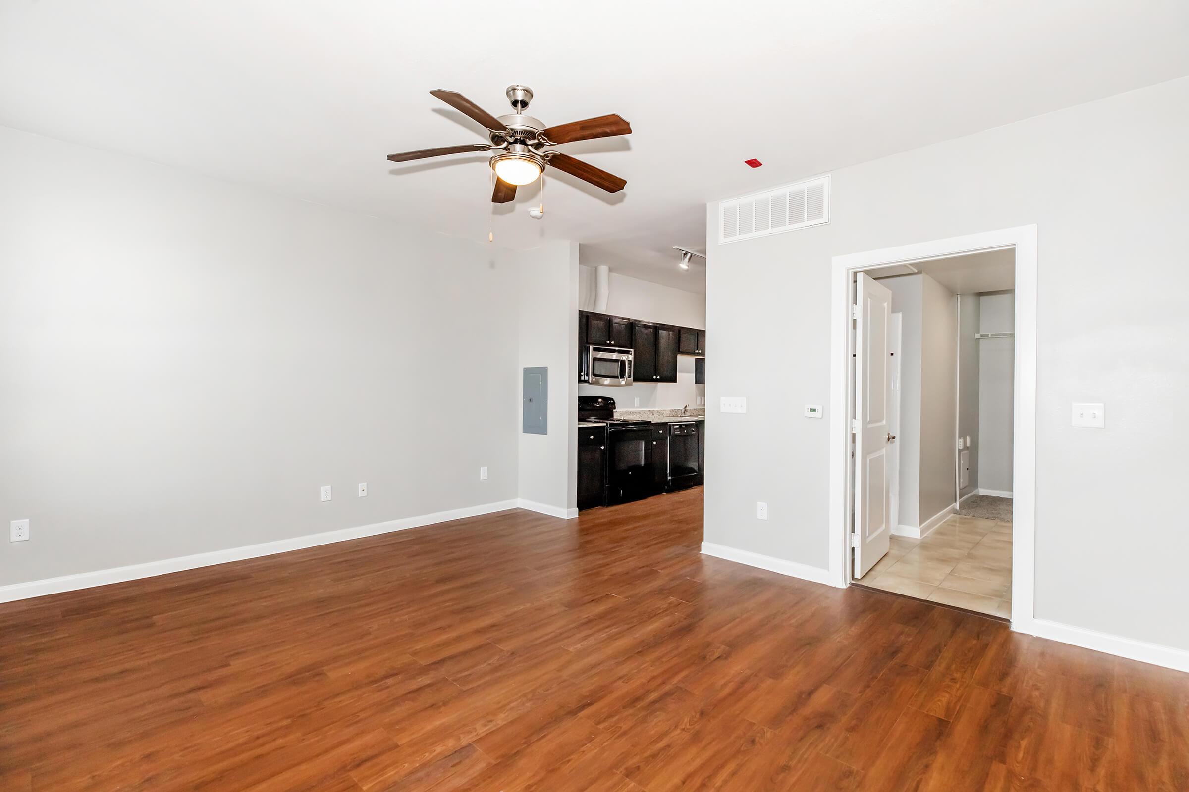 Spacious living area featuring a ceiling fan, dark cabinetry in the kitchen visible through an open layout, and a door leading to a small closet. Walls are painted light gray, and the floor is finished with wooden laminate, enhancing the modern and inviting atmosphere of the space.