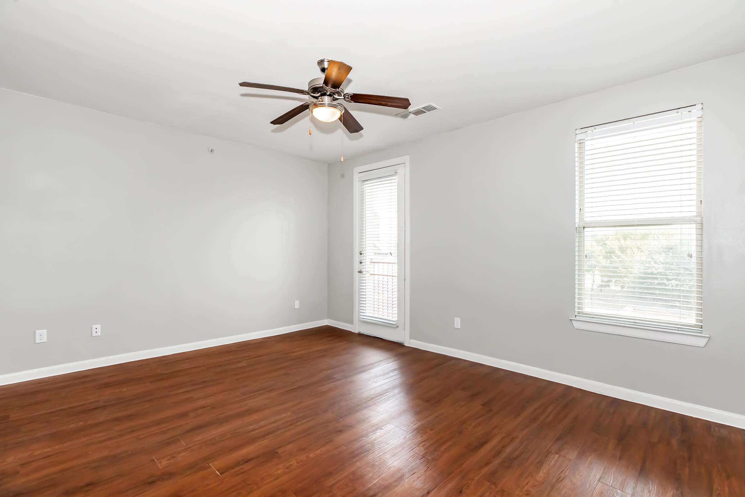 A spacious room with light gray walls, a ceiling fan, and hardwood flooring. One window with blinds allows natural light, and a door leads to a balcony or outdoor area. The room is empty, creating a bright and airy atmosphere, suitable for various uses such as a bedroom or living area.