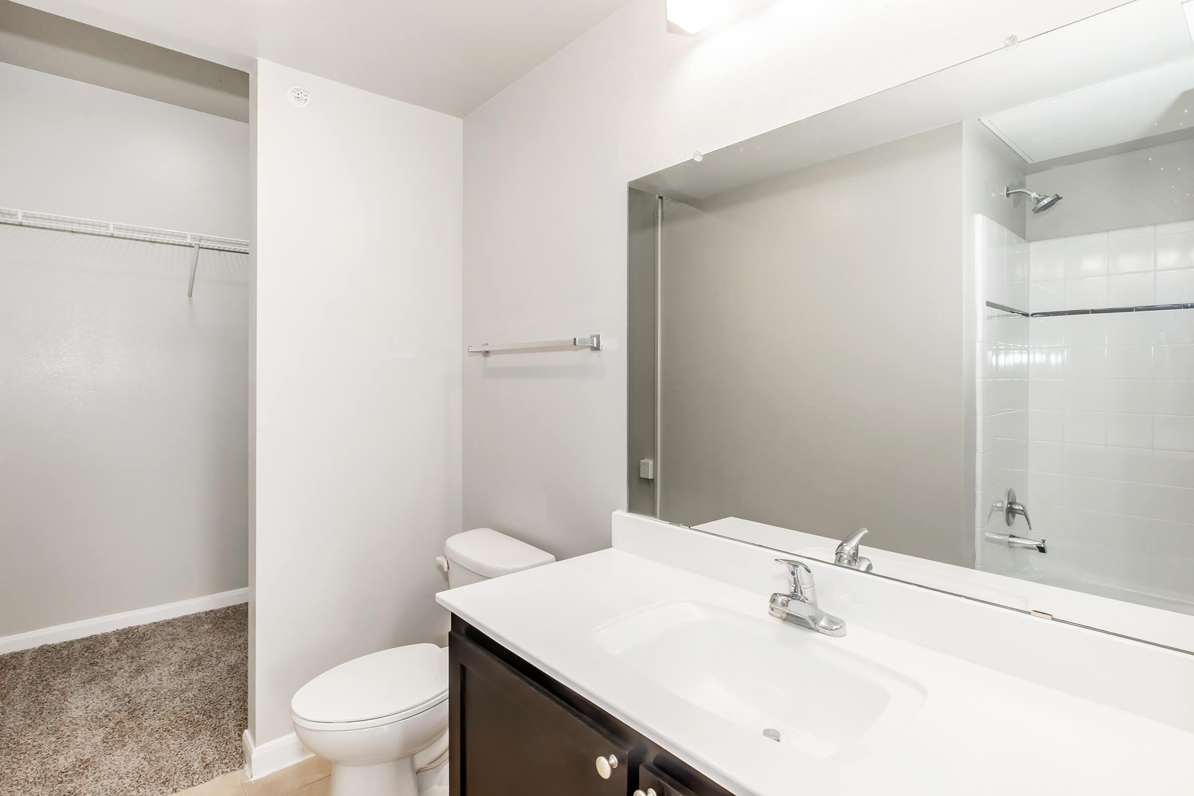 A clean, modern bathroom featuring a white countertop with a sink, a mirror above it, a toilet, and a spacious shower area. There is also a closet with a single shelf visible in the background, and the walls are painted in neutral tones. The floor is covered with light-colored carpet.