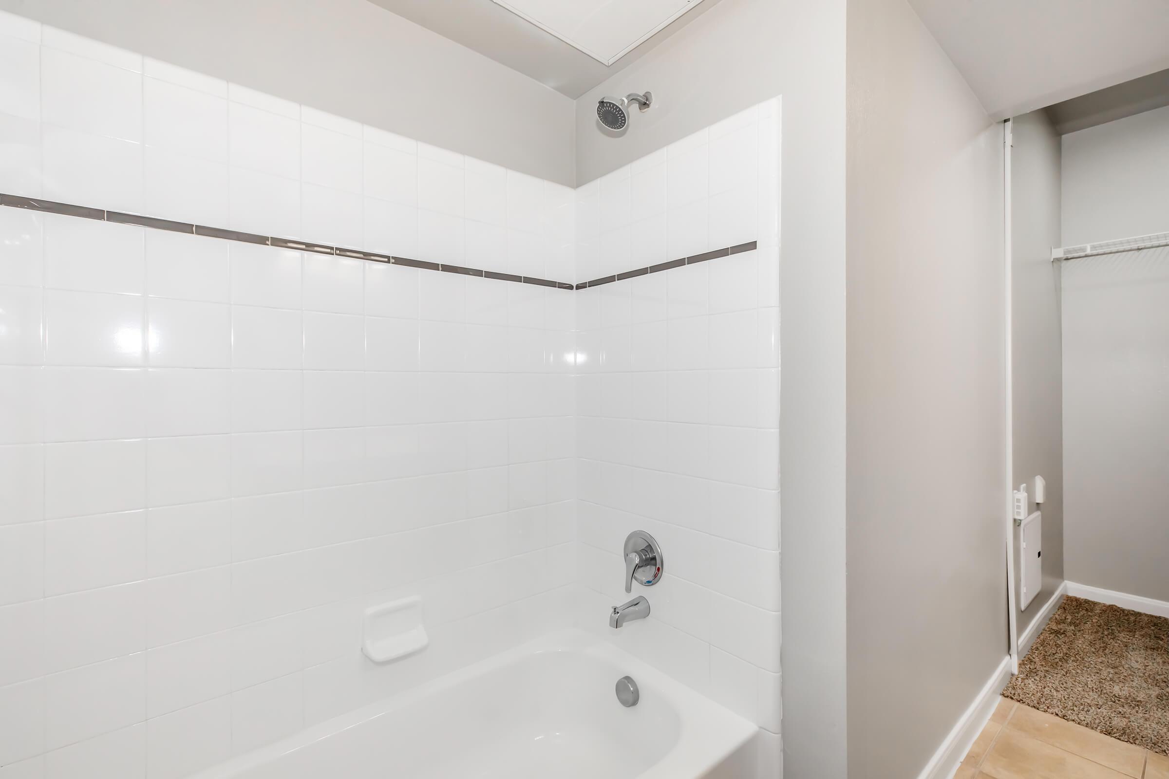 A clean, minimalistic bathroom featuring a white tiled shower and bathtub combination. The walls are painted in a light gray shade, and there is a small shelf above the shower area. A beige carpeted floor is visible in the adjacent room, adding a warm contrast to the tiles.