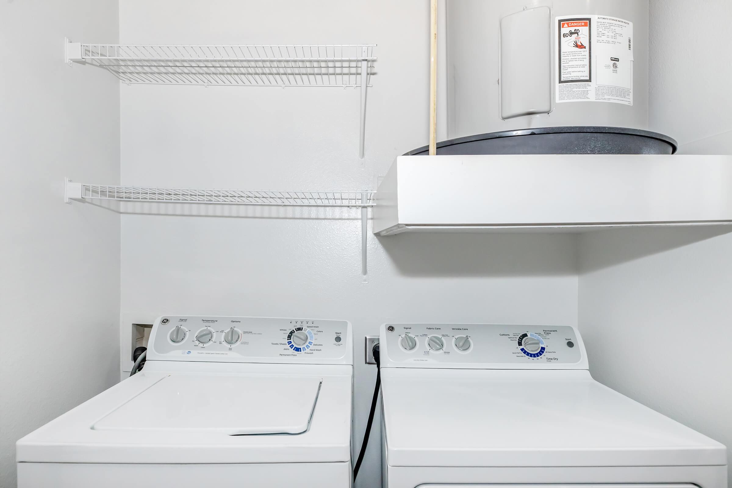 A clean laundry room featuring a white washing machine and dryer side by side. Above them are storage shelves and a wall-mounted container. The walls are painted white, creating a bright and tidy atmosphere.