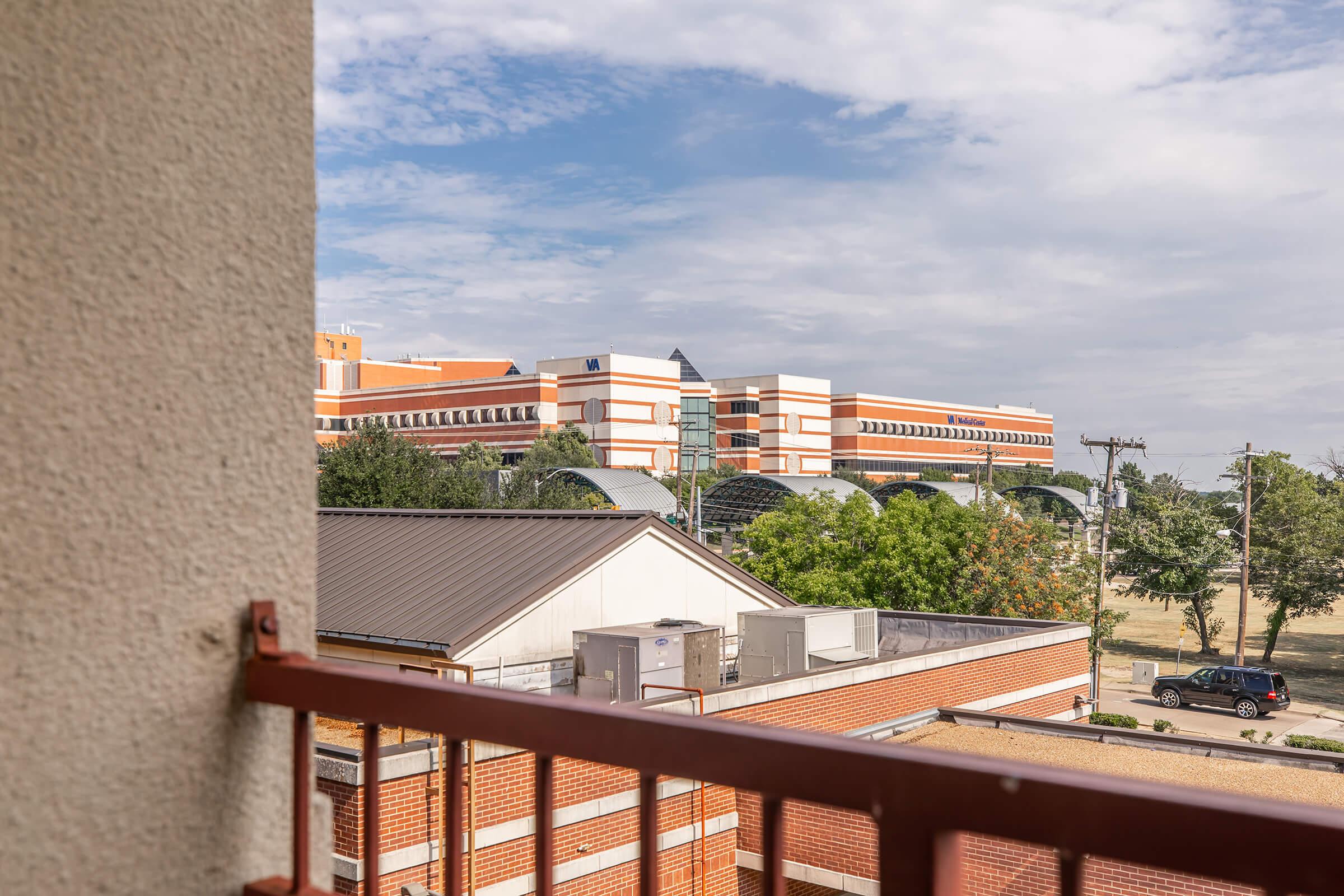 View from a balcony overlooking a multi-story building with a red and white brick facade. The sky is partly cloudy, and there are trees nearby. In the foreground, there is a metal railing and part of another building with air conditioning units.