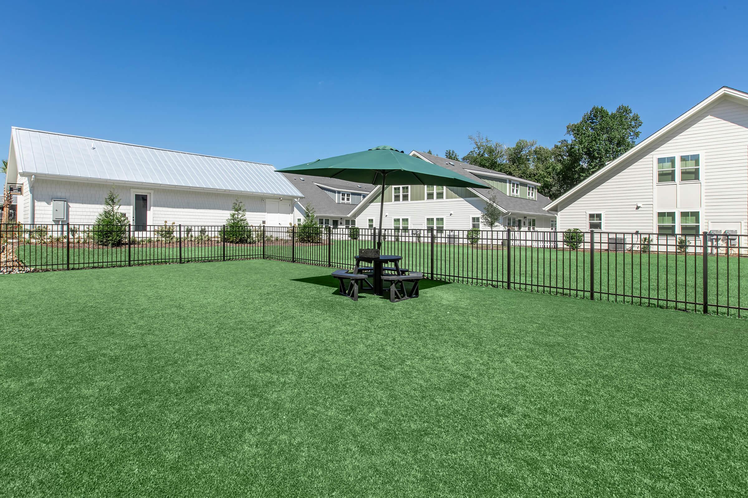 A green fenced-in yard featuring artificial turf, with a black picnic table and umbrella in the center. In the background, there are several white buildings under a blue sky with some trees lining the edges of the area. The scene conveys a clean and welcoming outdoor space.