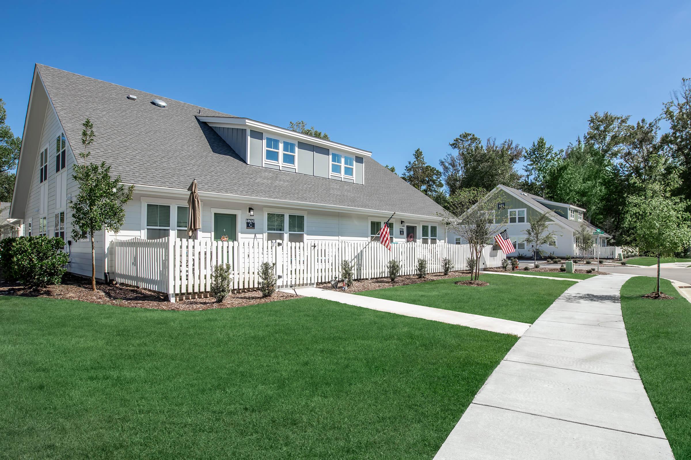A well-maintained residential area featuring several white houses with gray roofs, neatly landscaped lawns, and small trees. Sidewalks wind through the greenery, and American flags are displayed outside some of the homes. The scene is bright and sunny, with clear blue skies in the background.