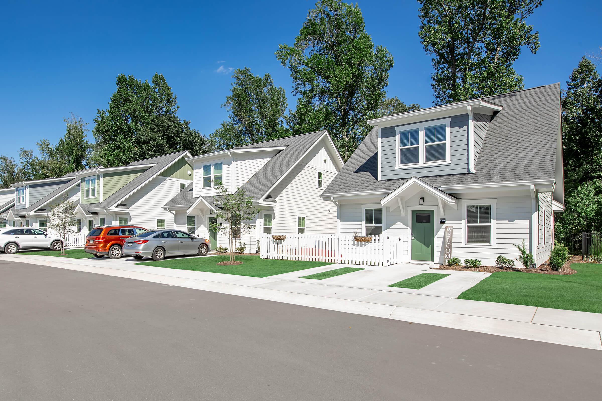 A row of modern single-family homes with light-colored siding and gabled roofs on a sunny day. Each house has a landscaped front yard and a driveway. Trees line the street, providing a natural backdrop to the neighborhood.