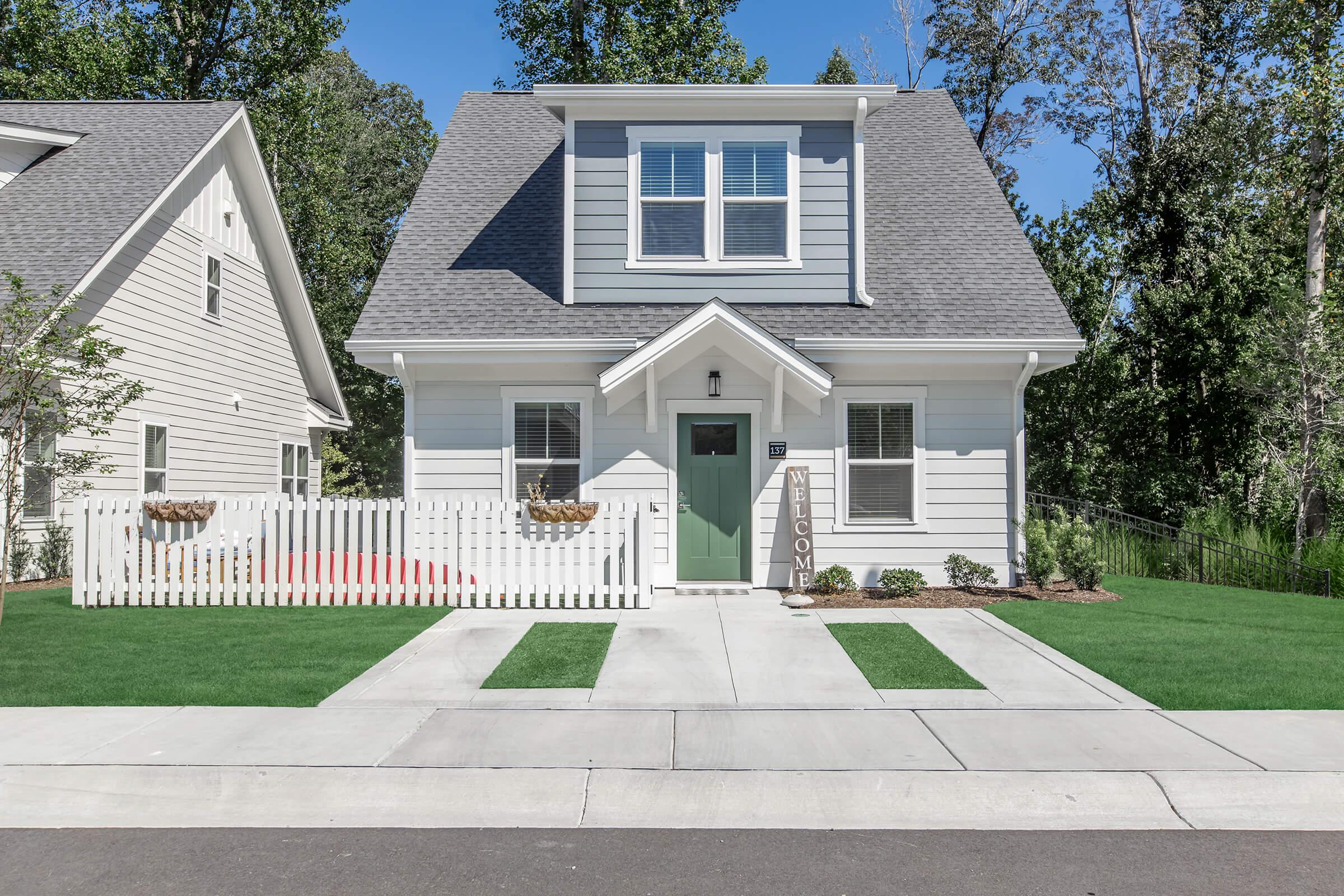 A two-story, modern white house with a green door, surrounded by landscaped grass and small plants. A white picket fence encloses the front yard, which has a "WELCOME" sign by the door. The house features a sloped roof and large windows, set against a backdrop of trees.