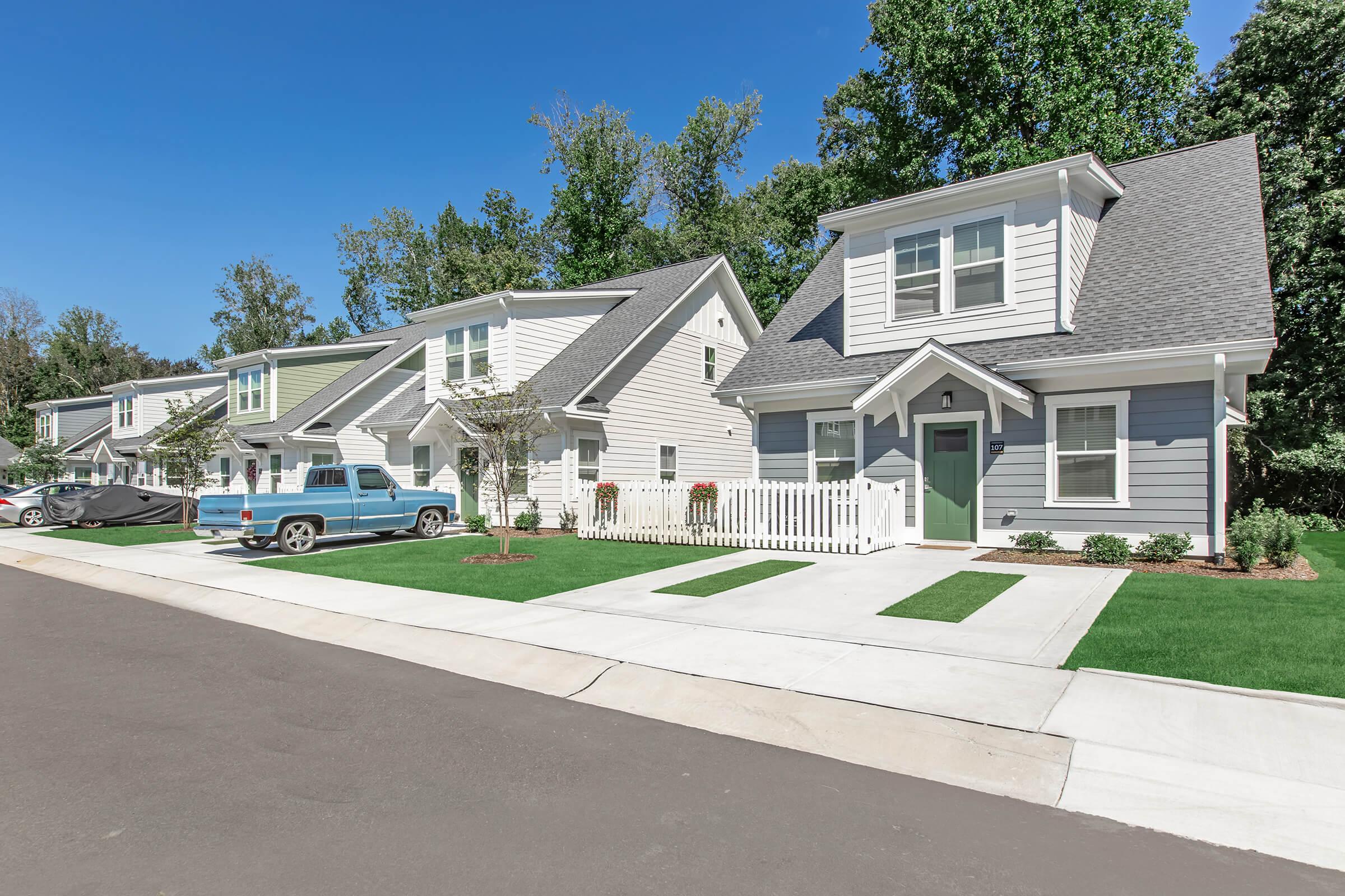 A row of modern suburban houses with light siding and dark roofs, featuring front porches and landscaped yards. A blue pickup truck is parked nearby on a paved street, with green trees in the background under a clear blue sky.