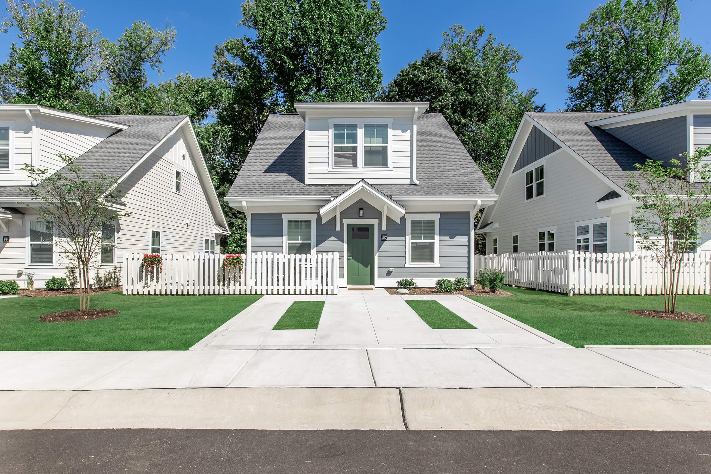 A charming two-story house with a gray exterior, white trim, and a welcoming front porch. The yard features well-maintained grass, bordered by a white picket fence. Two trees provide shade, and the surrounding neighborhood has similar styled homes, all under a clear blue sky.