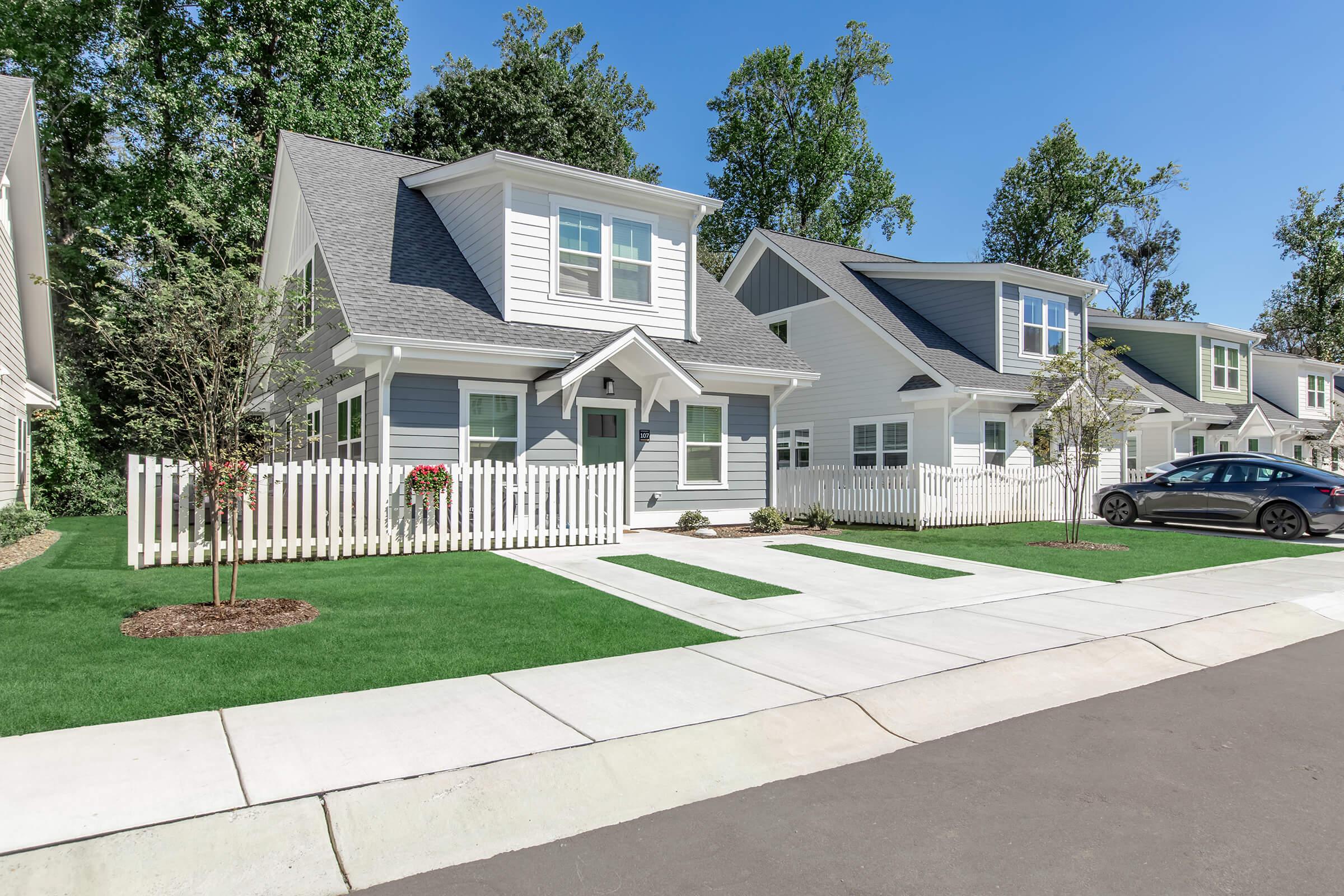 A suburban neighborhood featuring two modern homes with light gray siding and white trim. Each house has a well-maintained lawn, small trees, and white picket fences. A driveway leads up to the houses, with one car parked in front. The scene is set on a sunny day with clear blue skies.