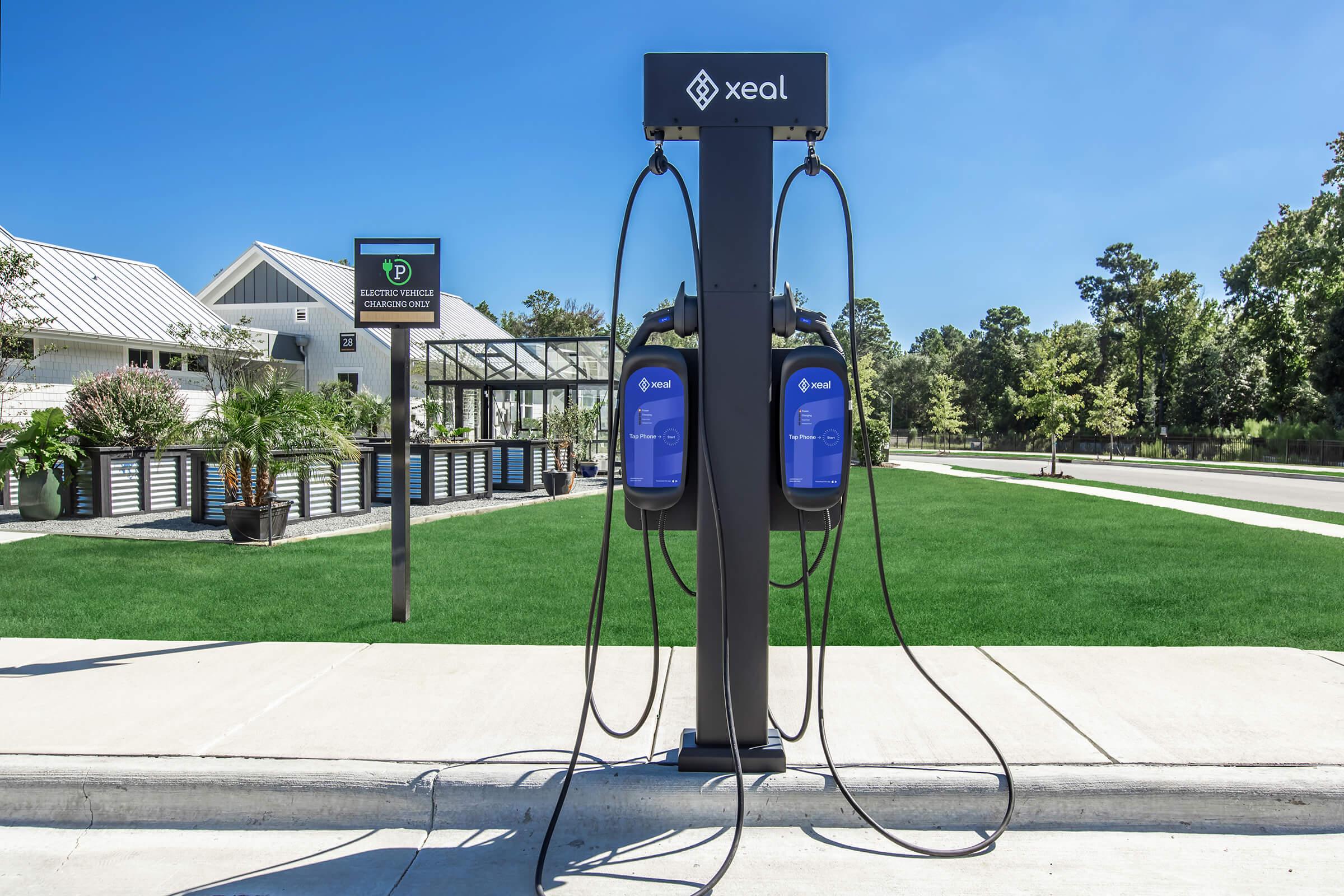 An electric vehicle charging station featuring two blue charging units mounted on a black post. The station is located on a paved area with green grass around it, and a sign in the background indicates the location as a charging point. A building is visible in the distance.