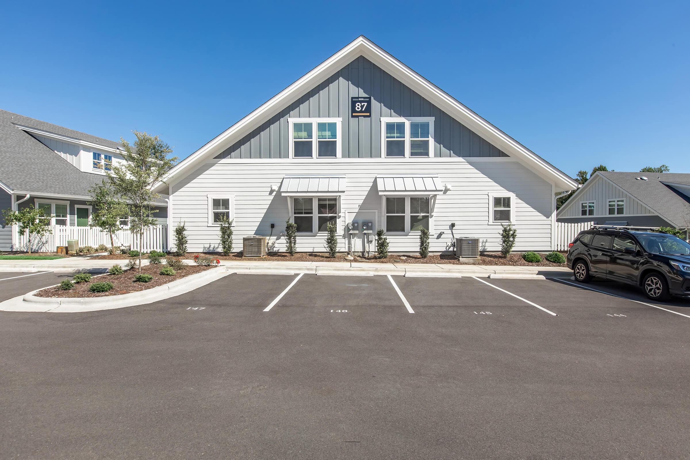 A modern, white two-story building with gray accents, featuring parking spaces in front. The building has large windows and awnings, and is surrounded by landscaped areas with small shrubs. Clear blue sky overhead, creating a bright and inviting atmosphere.