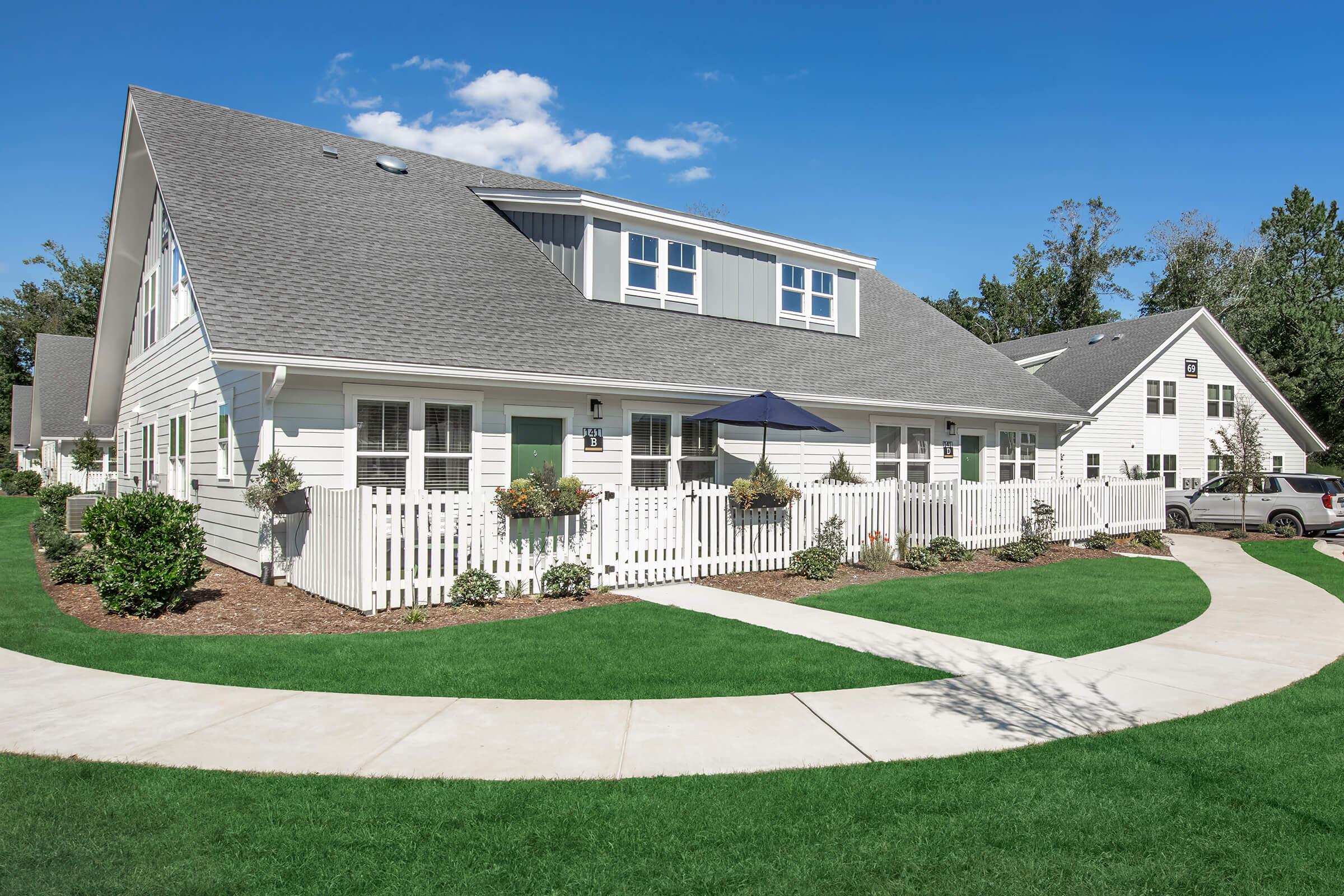 A well-maintained residential complex featuring white buildings with gray roofs. The landscape includes green lawns and neatly trimmed bushes, along with a white picket fence. Several walkways lead through the area, and there is an umbrella visible for outdoor seating. Bright blue sky completes the picturesque scene.