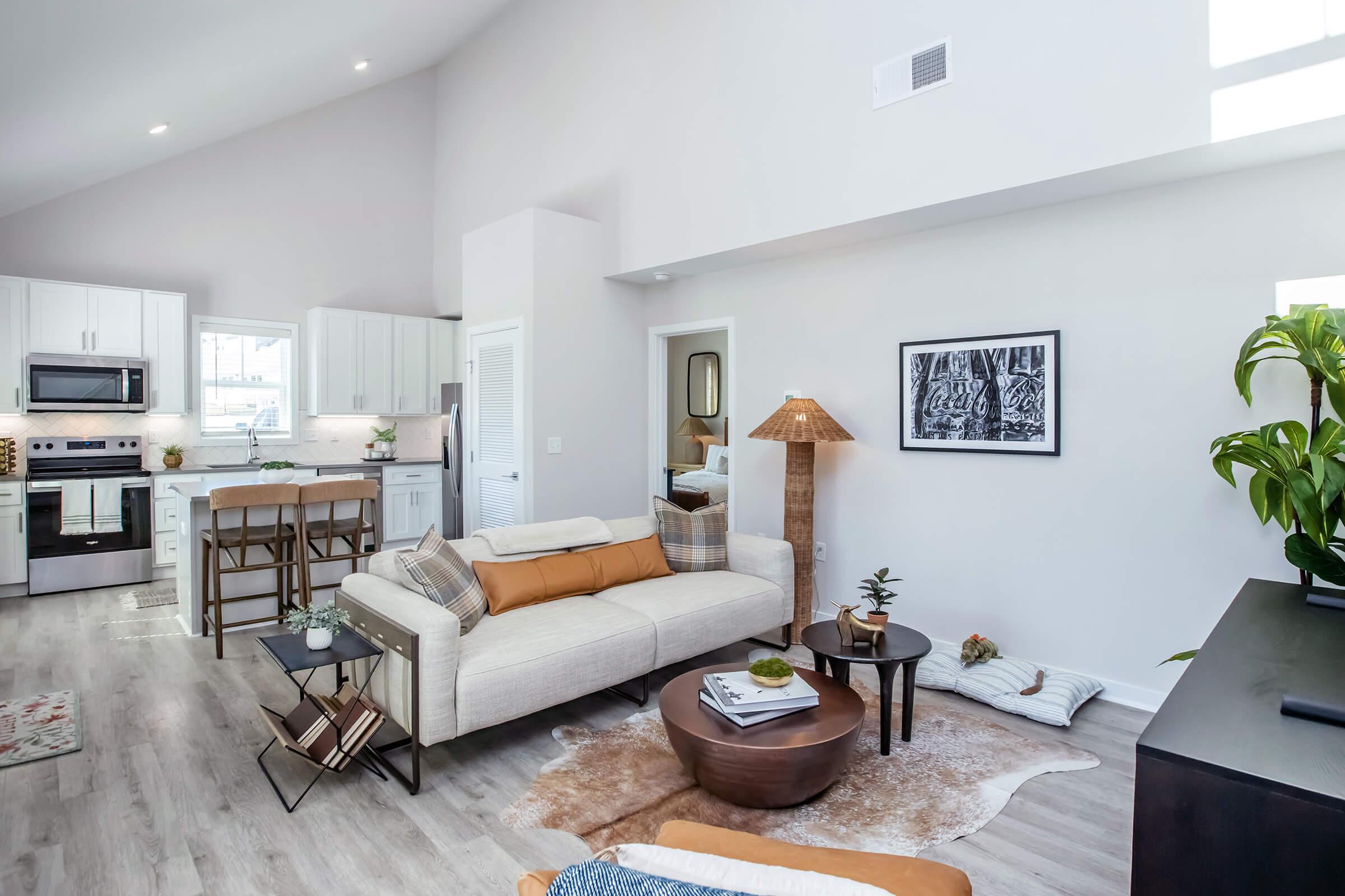 A modern living room featuring a light-colored sofa with orange and brown cushions, a round coffee table on a light rug, and a stylish floor lamp. In the background, a kitchen with white cabinets and stainless steel appliances is visible. Decorative plants and art add a touch of warmth to the space.