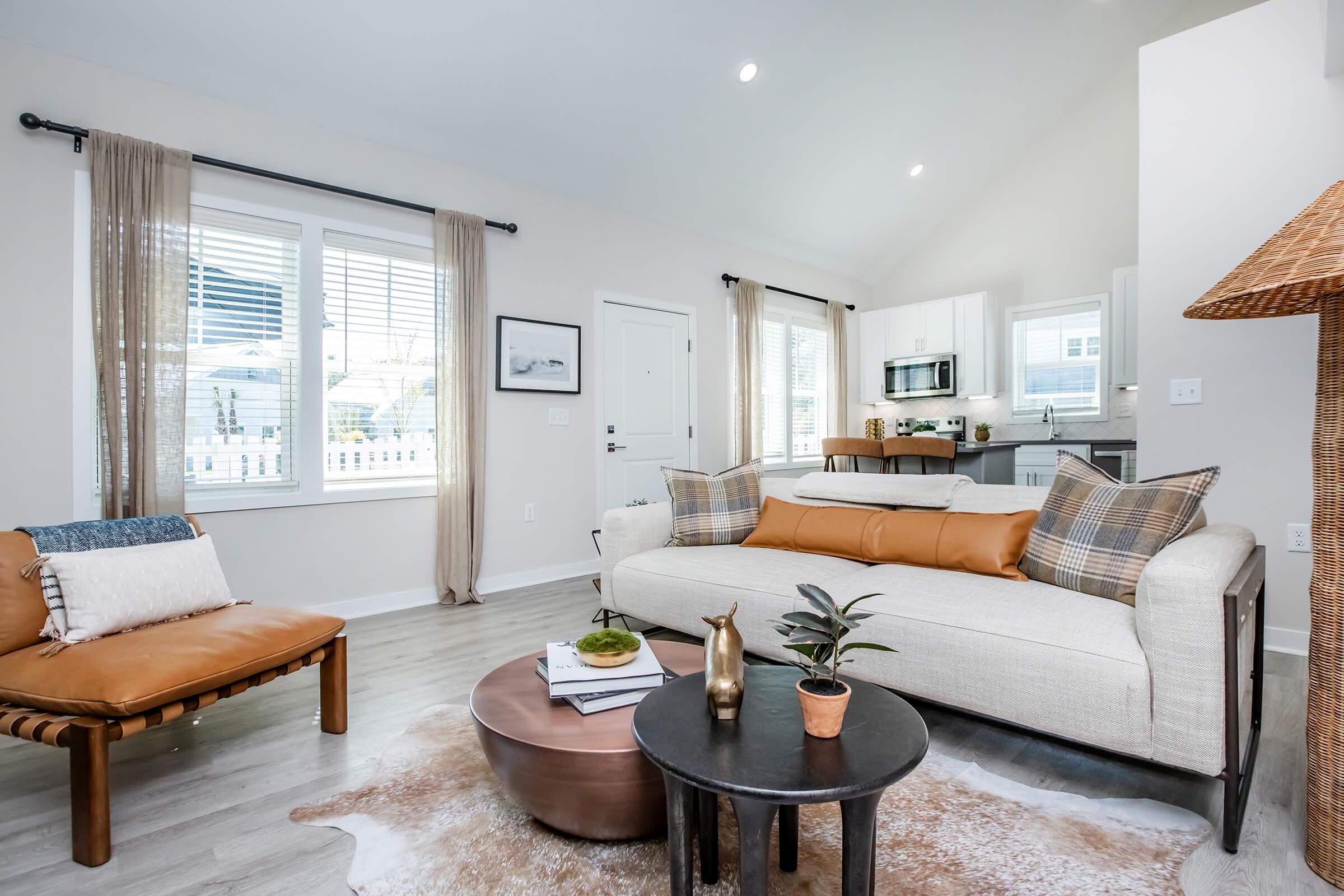 A modern living room featuring a beige sofa with plaid cushions, a leather accent chair, and a round coffee table on a cowhide rug. Natural light floods the space through large windows with sheer curtains. In the background, a stylish kitchen can be seen, enhancing the cozy atmosphere.
