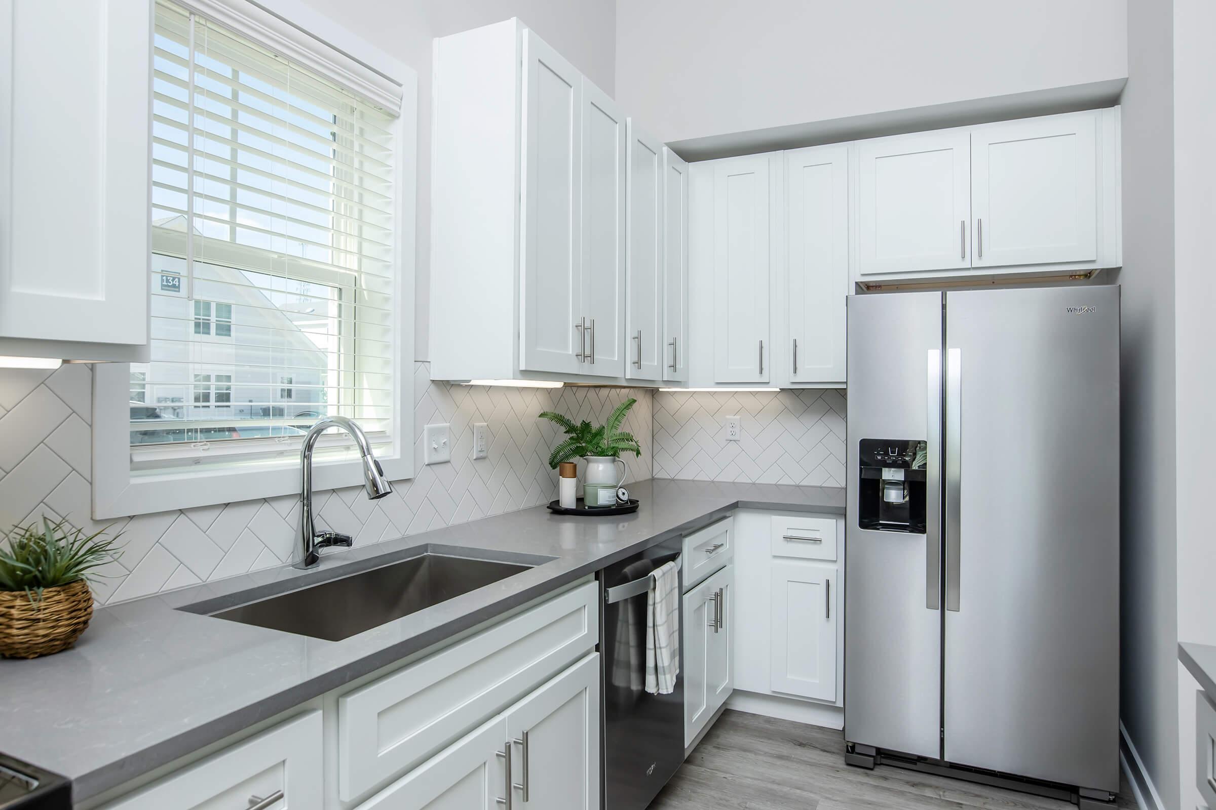 Modern kitchen featuring white cabinetry, a stainless steel refrigerator, and a sleek gray countertop. A large window allows natural light to illuminate the space, with a small potted plant on the countertop for a touch of greenery. The design includes a minimalist, clean aesthetic with a herringbone tile backsplash.