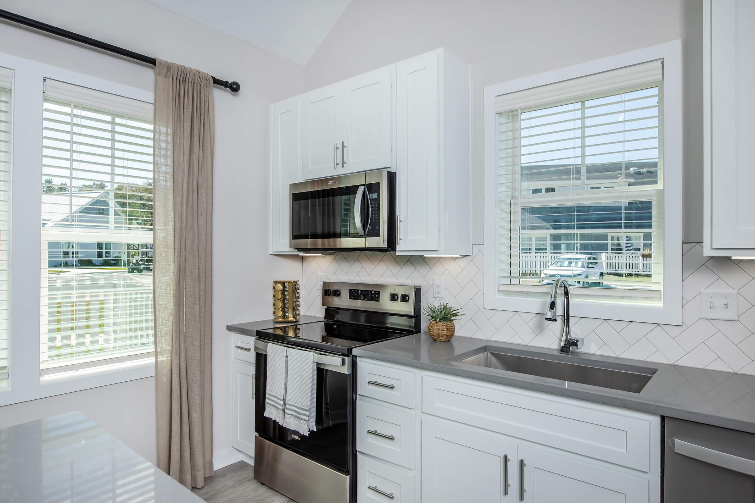 Modern kitchen featuring white cabinetry, stainless steel appliances including a microwave and an oven, a gray countertop, and a sink. Natural light streams through two windows with white blinds. Decorative elements include a small potted plant and a gold vase, enhancing the contemporary design.