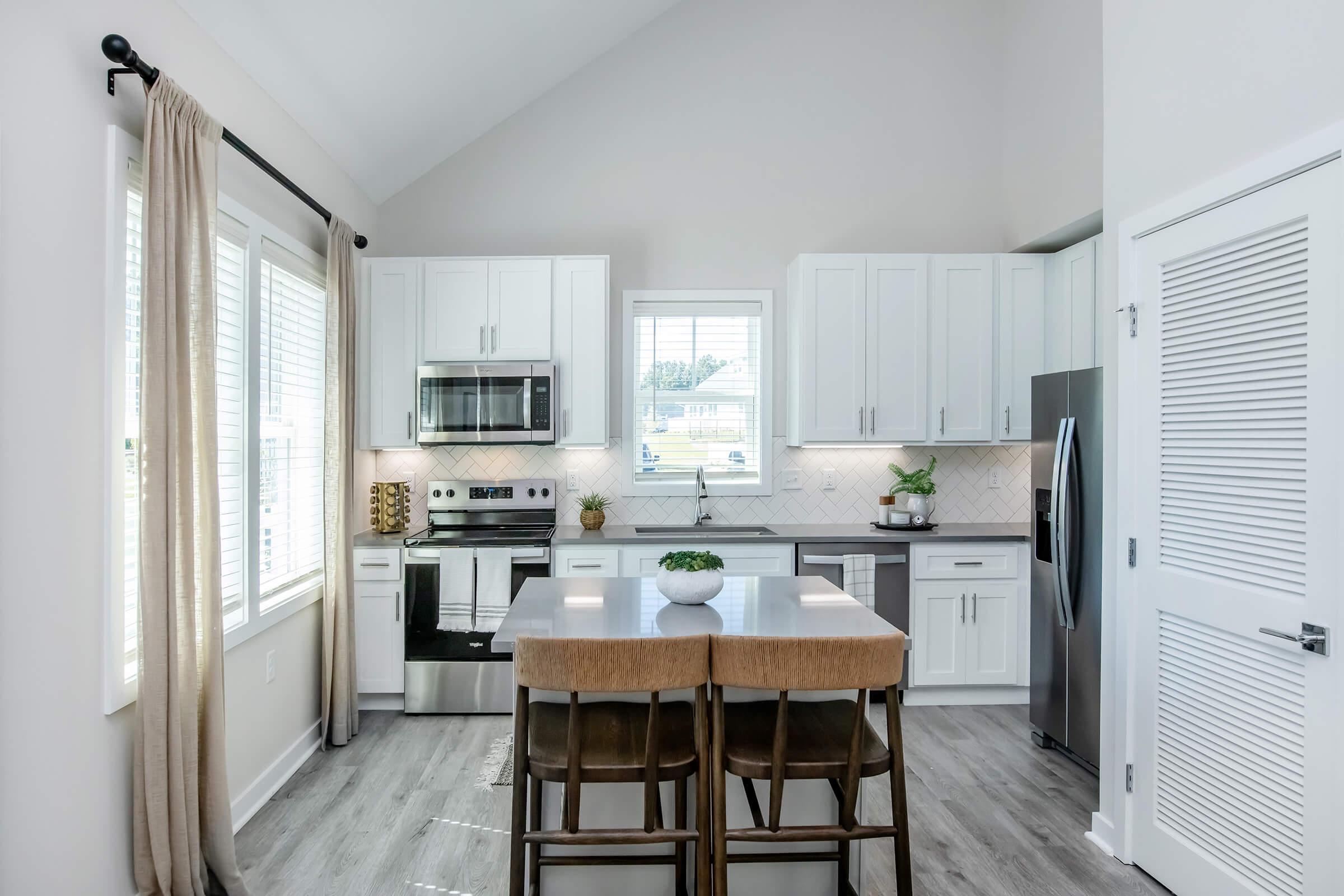 Modern kitchen featuring white cabinets, stainless steel appliances, and a large island with seating for three. Natural light floods in through large windows, highlighting the light gray walls and stylish decor elements, creating a bright and inviting atmosphere.