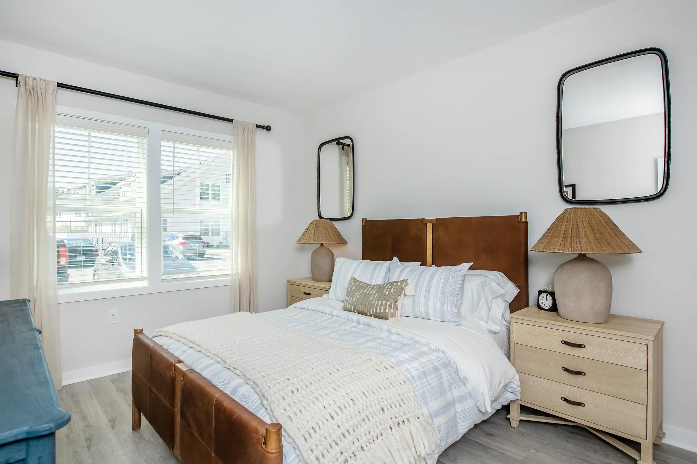 A cozy bedroom featuring a queen-sized bed with a leather headboard, two bedside tables with decorative lamps, and a woven blanket. The room has large windows with sheer curtains, allowing natural light to fill the space, and features modern mirrors on the walls.