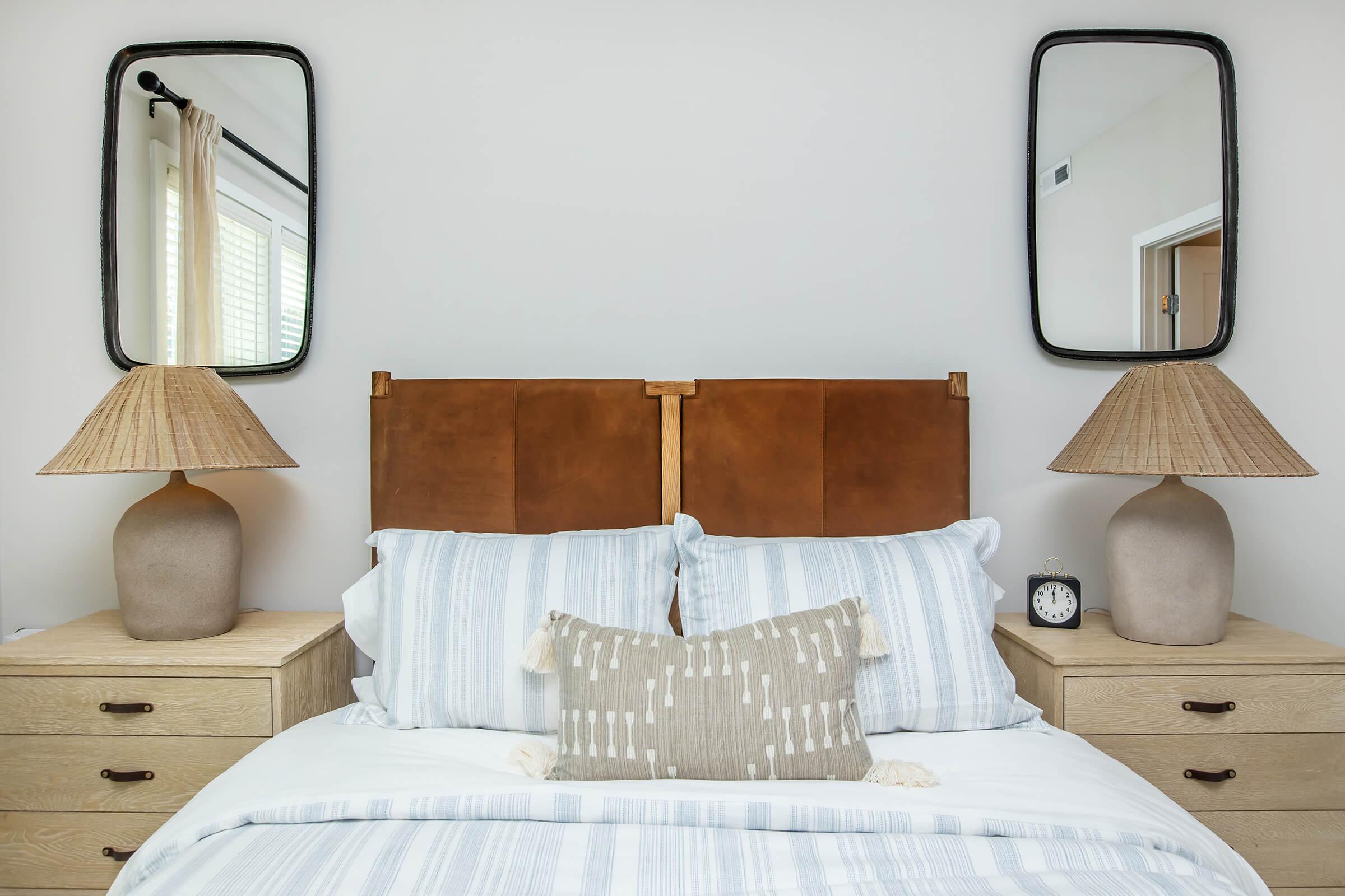 A cozy bedroom featuring a brown leather headboard, light blue striped bedding, and decorative pillows. On either side of the bed are wooden nightstands with textured lamps and mirrors above. A small clock sits on one nightstand, adding a touch of elegance to the serene atmosphere.
