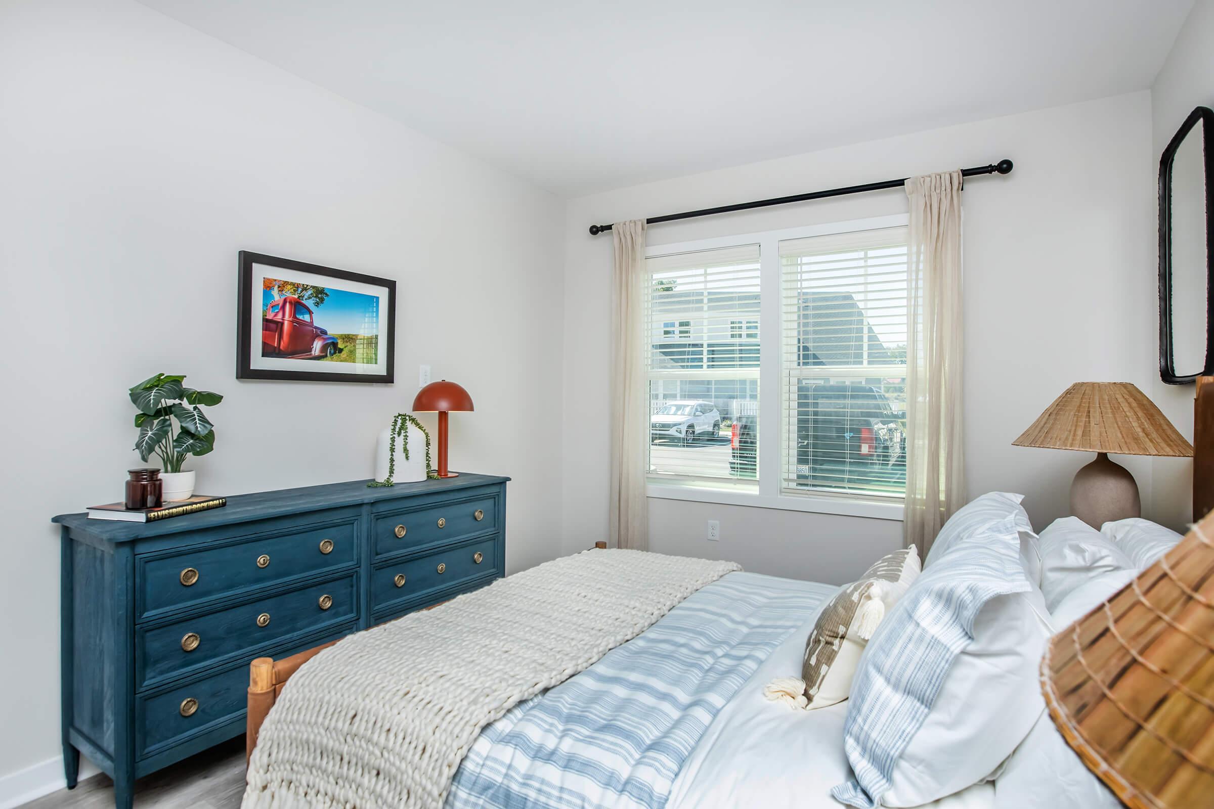 A cozy bedroom featuring a blue dresser, neatly made bed with striped pillows, and decorative elements like a lamp and indoor plant. Natural light streams through a window with sheer curtains, complemented by a framed picture on the wall. The overall decor is modern and inviting.
