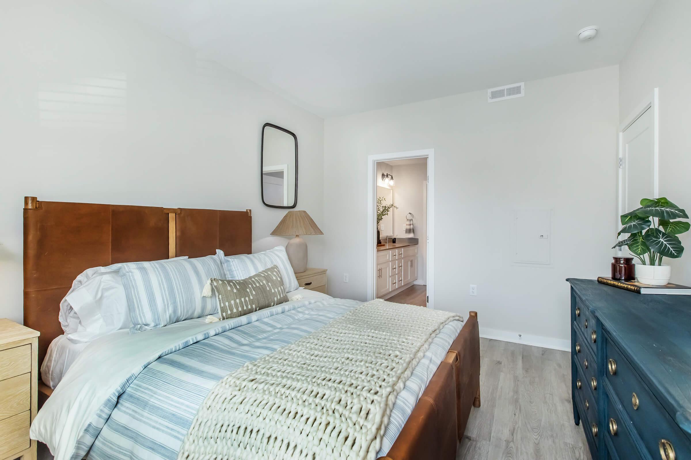 A cozy bedroom featuring a brown leather bed with a white and blue striped blanket and decorative pillows. There’s a bedside lamp and a wooden dresser. In the background, a doorway leads to a well-lit area, possibly a bathroom, complemented by a potted plant on the dresser. The walls are painted in soft neutral tones.