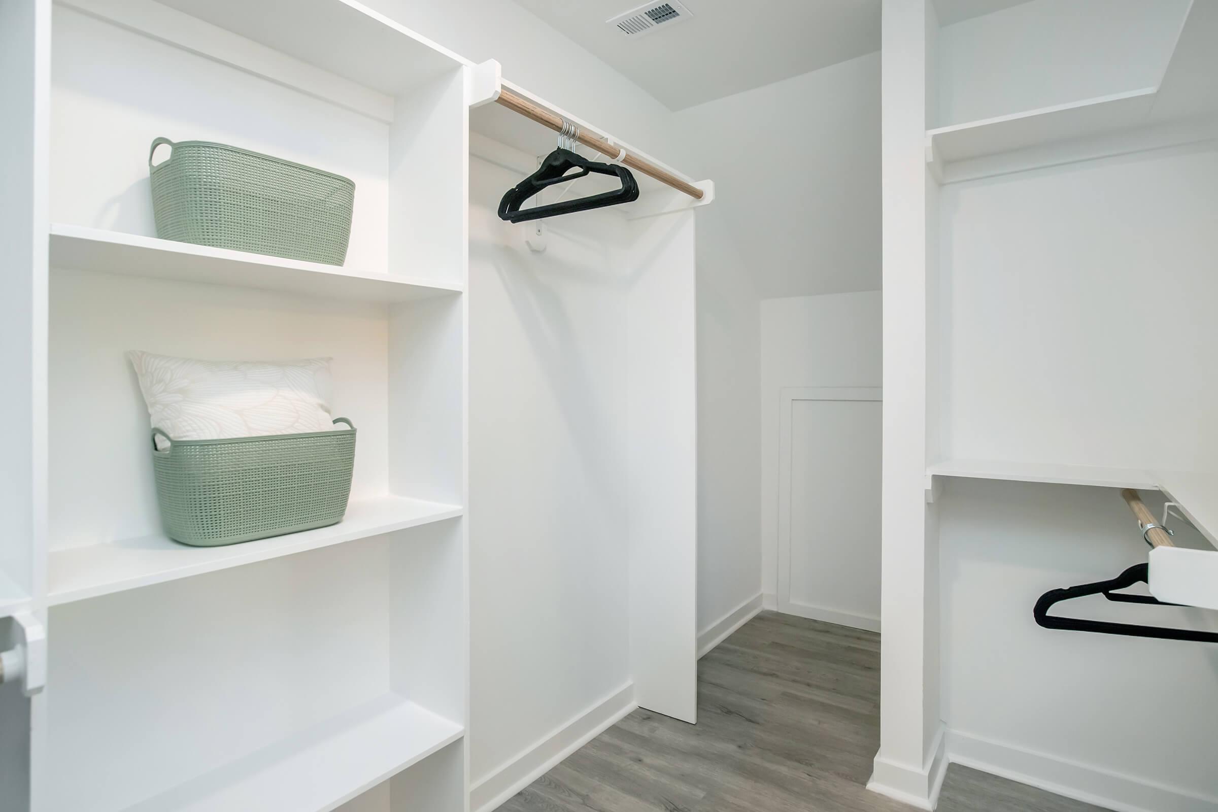 A well-organized closet featuring white shelves, a clothing rod with black hangers, and two green storage baskets. The floor has a light wood finish, and the walls are painted white, creating a clean and modern look.