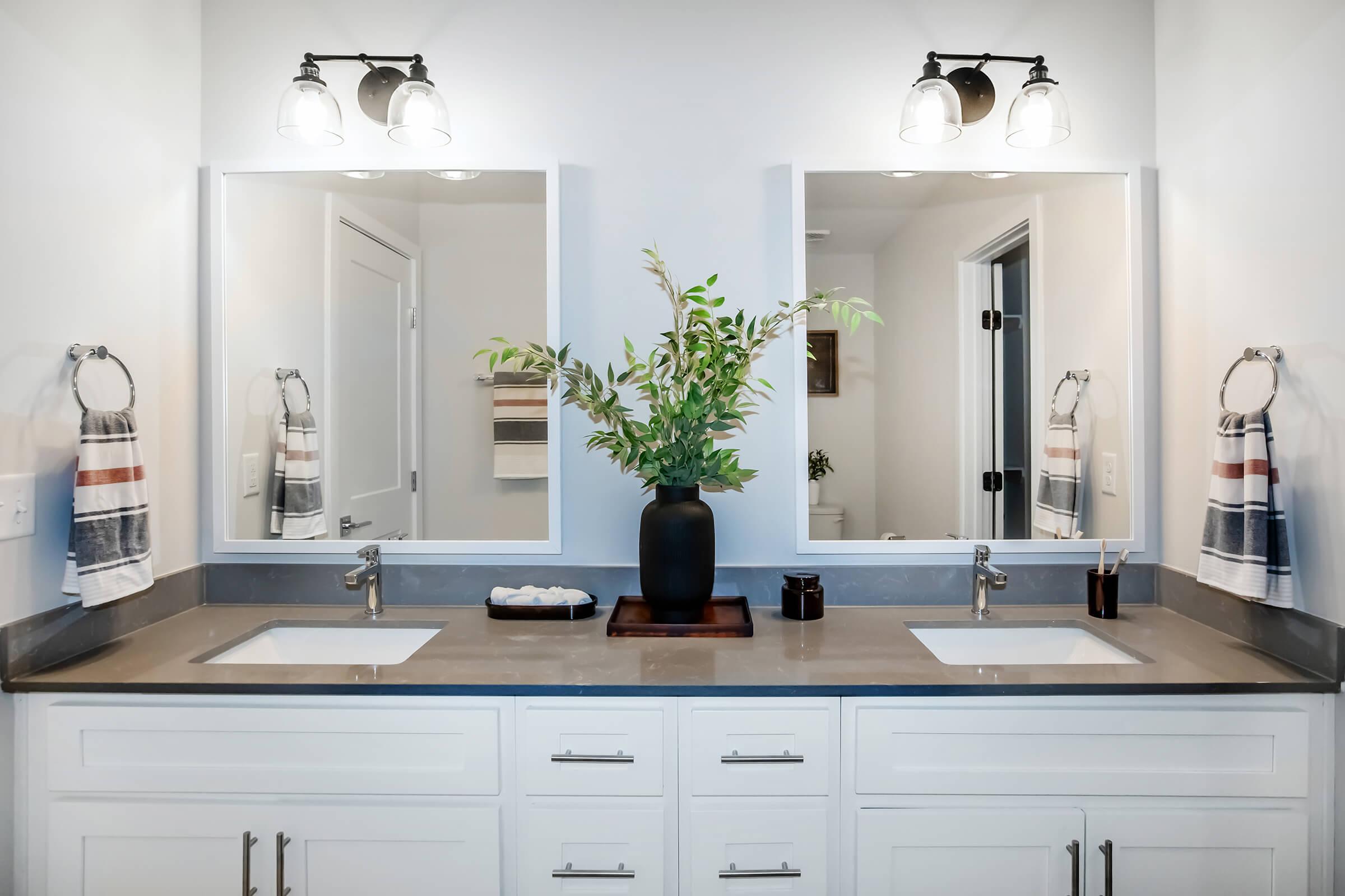 A modern bathroom with double sinks, two mirrors, and stylish lighting fixtures. The countertop is decorated with a black vase containing greenery, a small tray with decorative items, and hand towels hanging by each sink. The overall design is clean and contemporary.