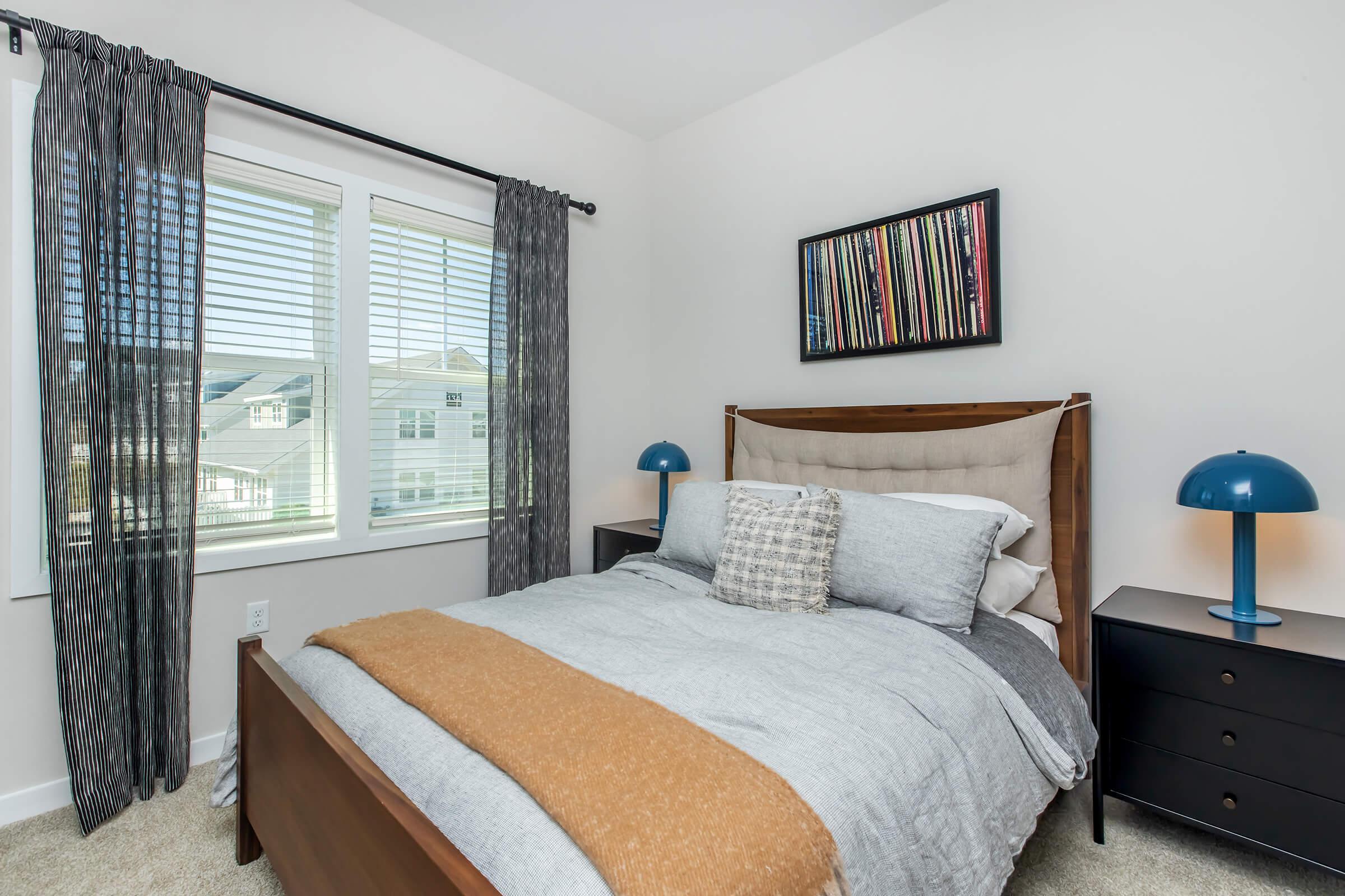 A cozy bedroom featuring a wooden bed with gray bedding and an orange throw. There are two bedside tables with blue lamps, and a large window with light-colored curtains allows natural light to fill the room. A framed artwork hangs above the bed, adding a touch of color to the neutral decor.