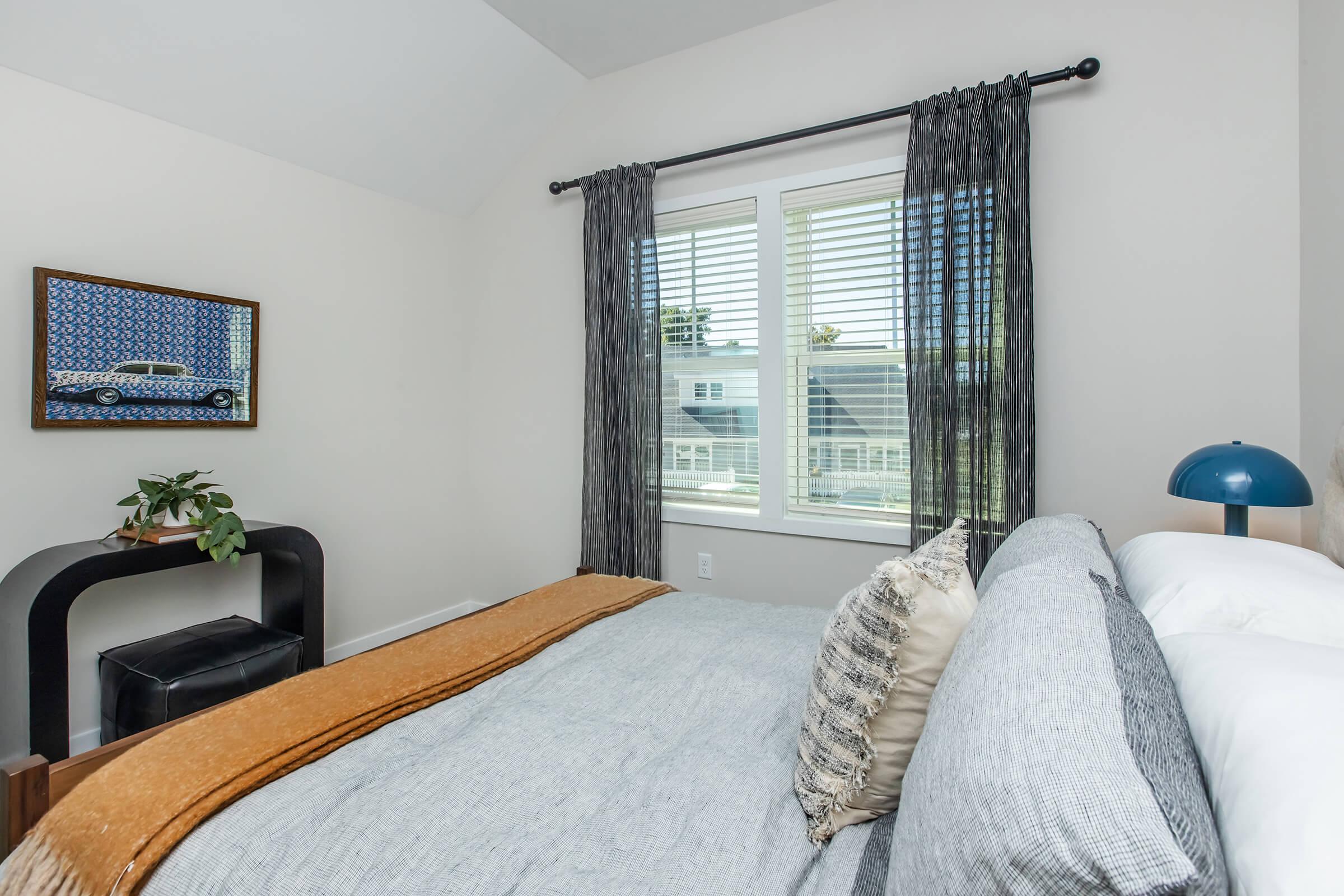 A well-decorated bedroom featuring a light gray bed with a textured blanket, two decorative pillows, and a bedside lamp. A window with blinds allows natural light in, and a small potted plant sits on a modern black nightstand. A framed picture hangs on the wall, depicting a vintage car.