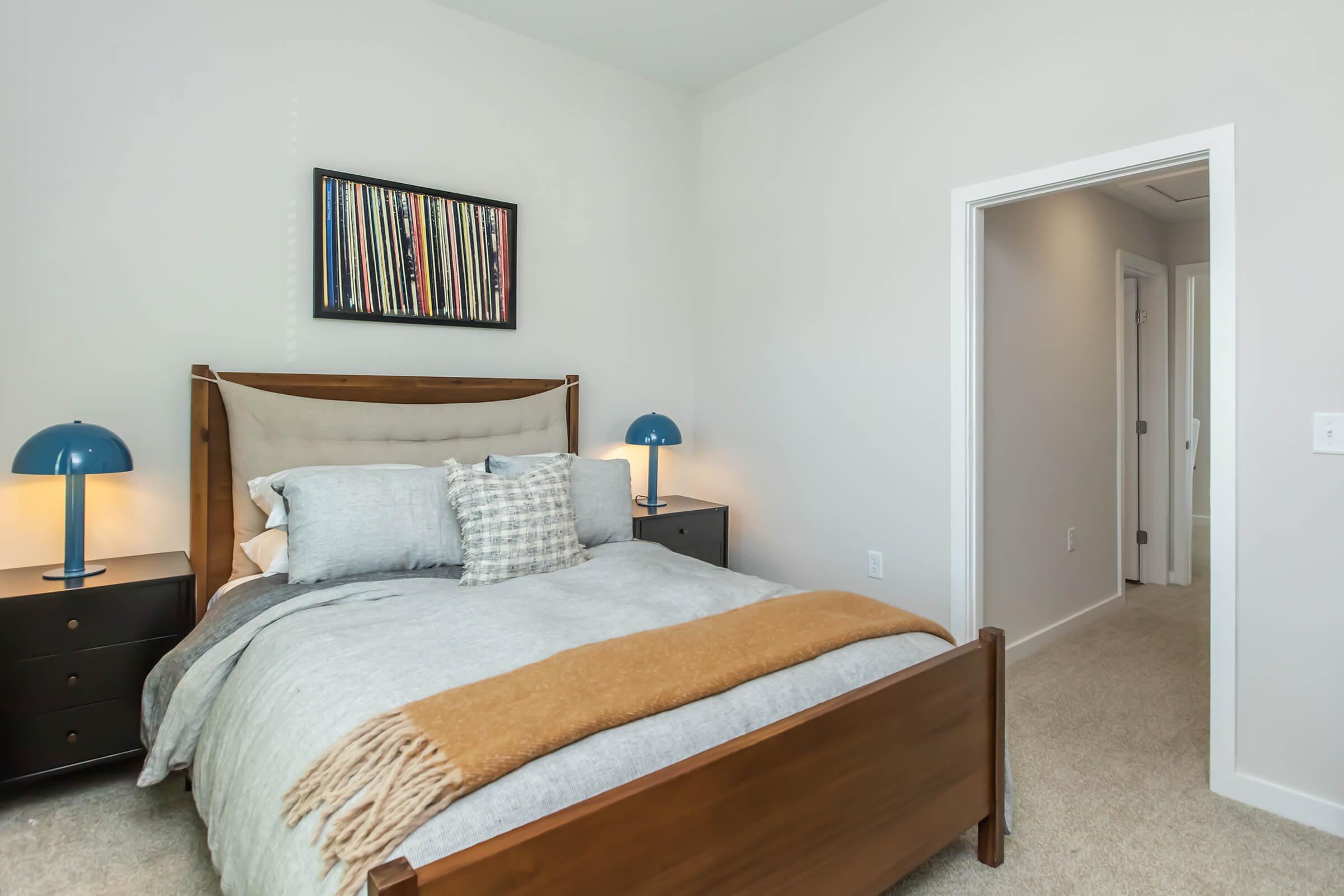 Cozy bedroom featuring a wooden bed with gray bedding and decorative pillows, two bedside tables with blue lamps, and a wall with framed artwork. A doorway leads to a hallway, and the room has light-colored walls and carpet.