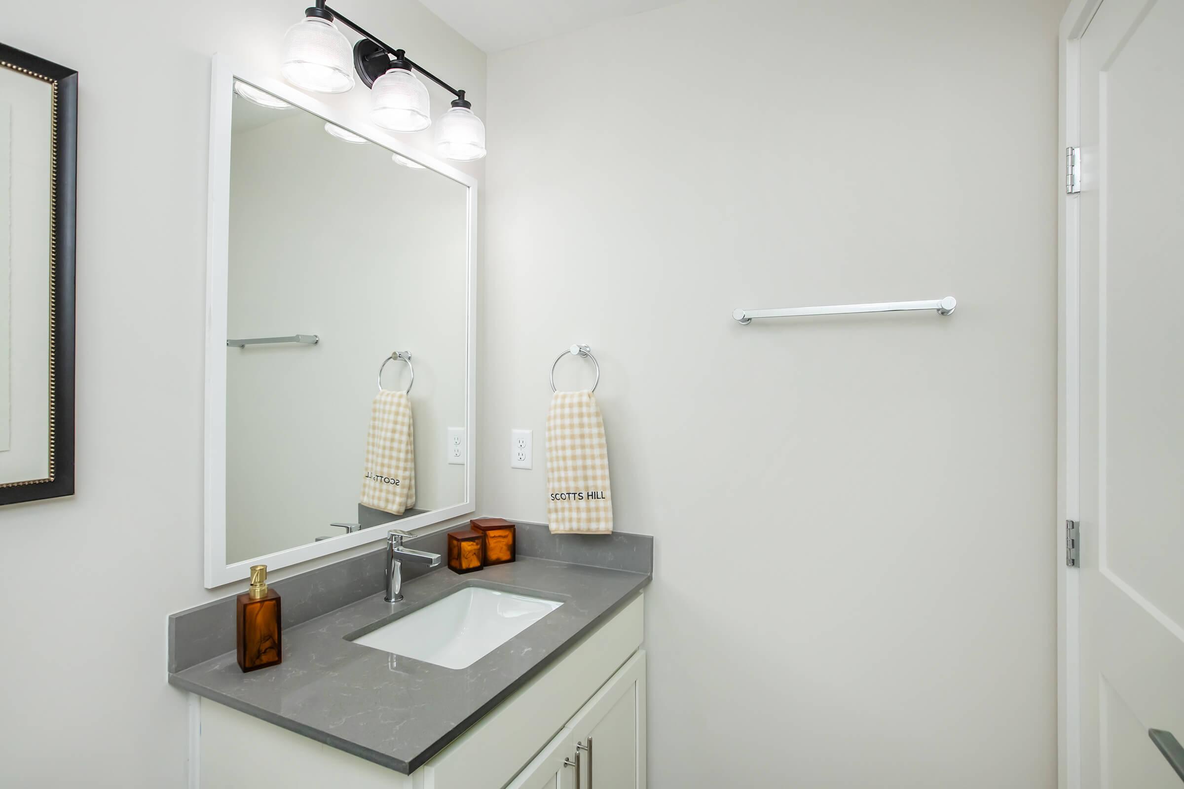 A well-lit bathroom featuring a modern gray countertop with a rectangular sink, a large mirror above, and two neatly hung hand towels. There are two amber-colored decorative bottles on the counter, and a towel bar in the background, all set against light-colored walls for a clean and airy look.