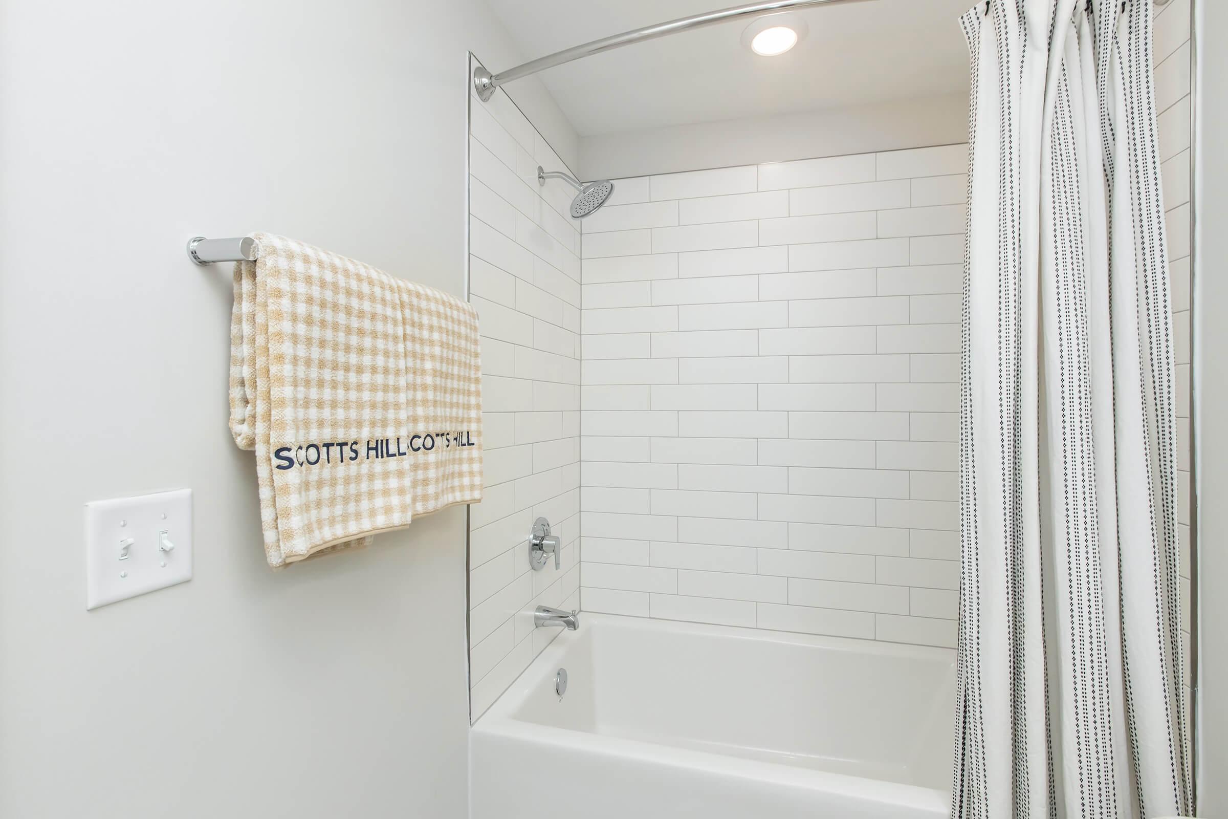 A clean bathroom featuring a white tiled shower area, a bathtub with a showerhead, and a towel rack holding a yellow checkered towel with "SCOTT'S HILL COASTAL" embroidered on it. The walls are light and minimalist, creating a bright and airy atmosphere.