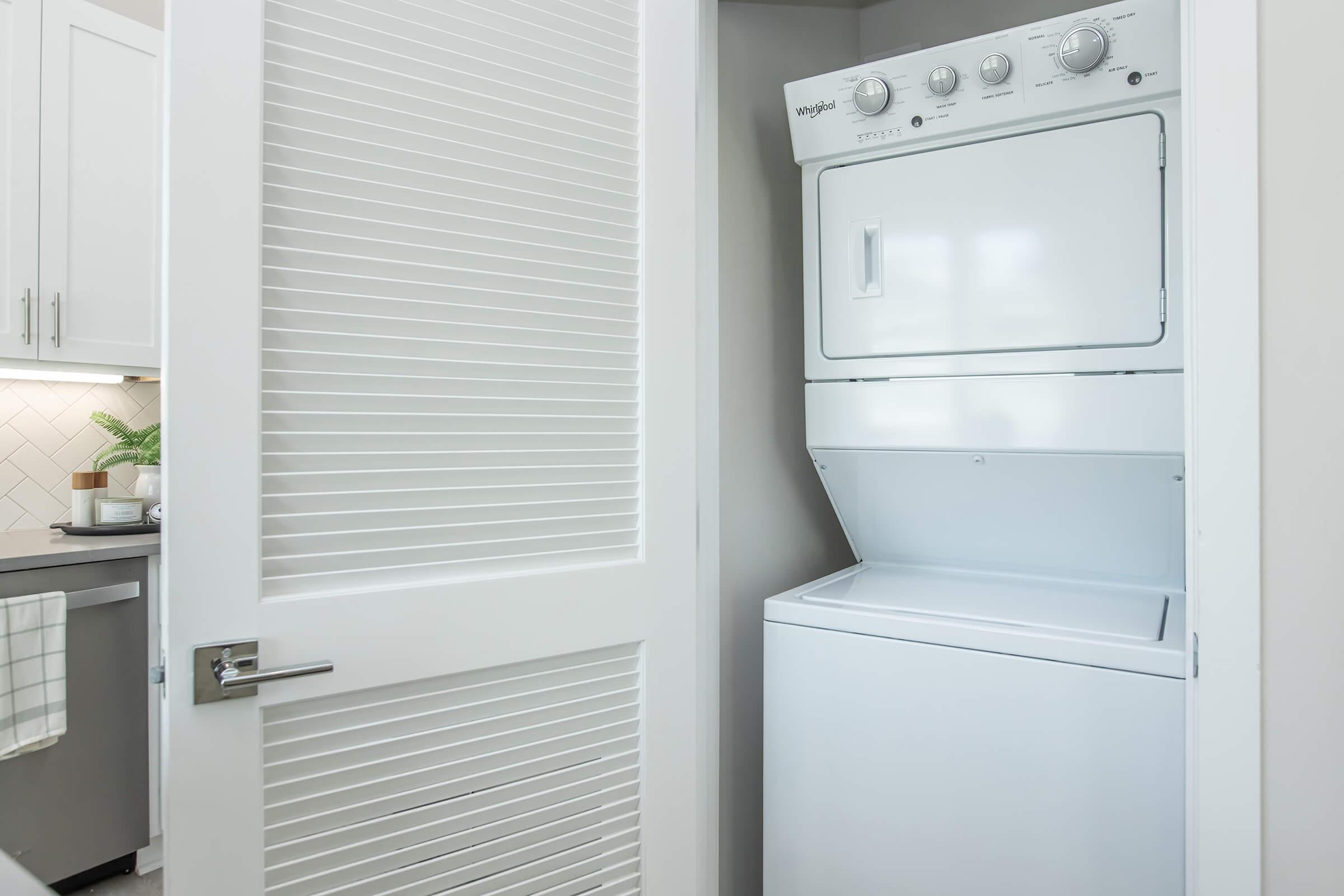 A white stacked washer and dryer unit is situated in a compact laundry space, partially hidden behind a closed white louvered door. The surrounding area features modern cabinetry and a tiled backsplash, contributing to a clean and organized home environment.
