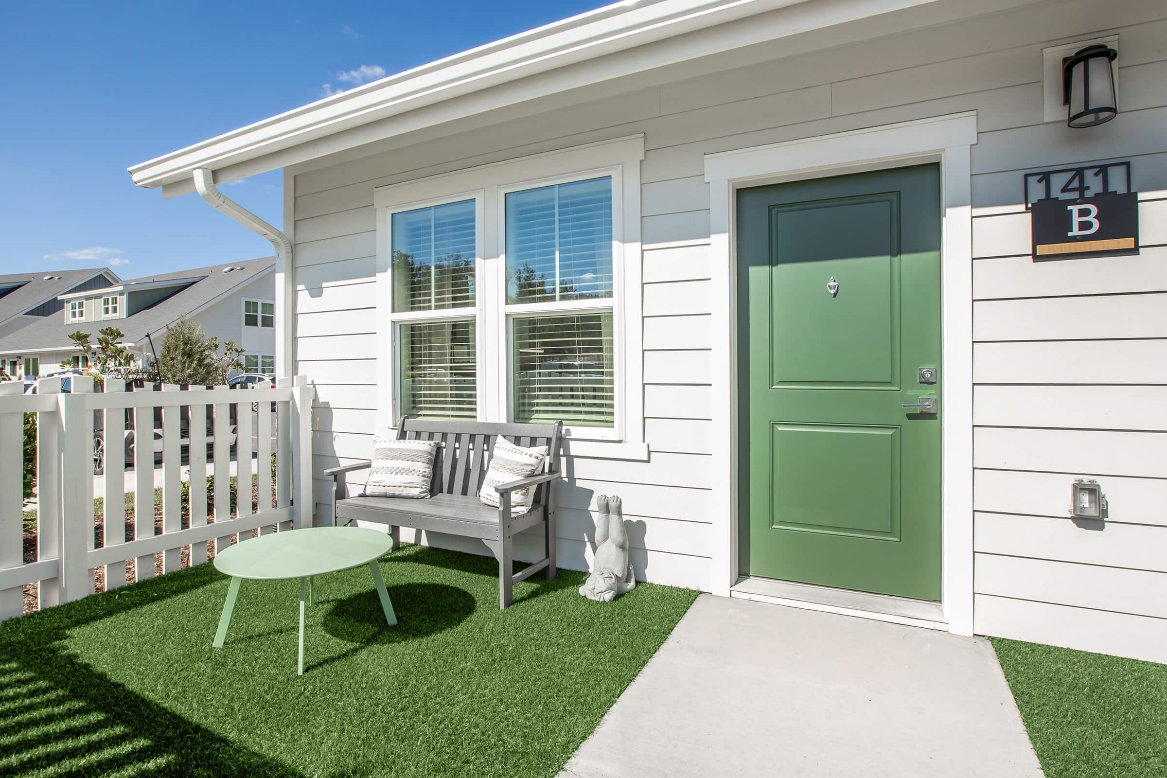 A cozy entryway featuring a green door with the number 141B displayed on the side. A small bench sits beside the door with a decorative pillow, and a light green side table is on the artificial turf. A small rabbit sculpture adds a charming touch to the welcoming outdoor space.