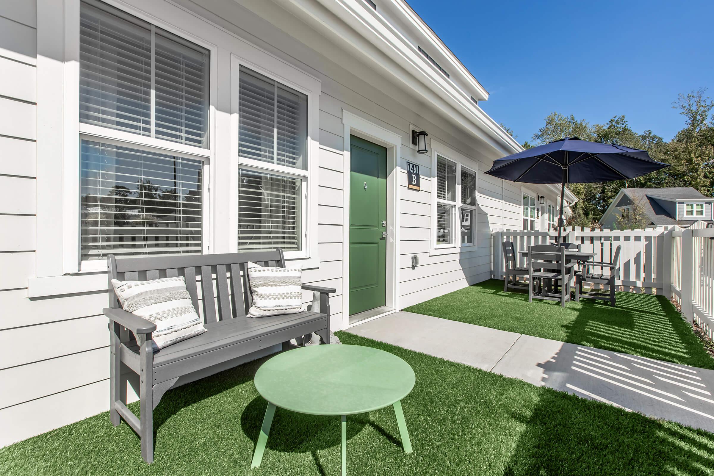 A small outdoor seating area featuring a gray bench and a round green table on faux grass. There is a large umbrella nearby, providing shade, and a white picket fence surrounds the space. The exterior wall of a house can be seen, with a door and windows allowing natural light.