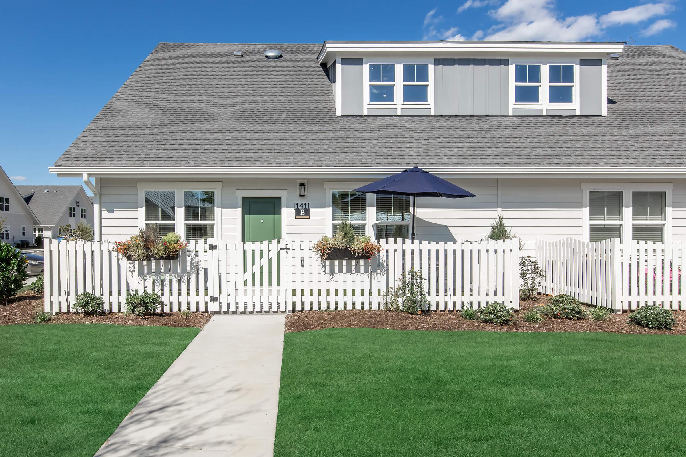 A charming two-story house with a grey roof and white exterior. The front yard features neatly trimmed grass, flower boxes with plants, and a white picket fence. A sidewalk leads to the entrance, which has a green door and a small umbrella beside it, providing shade. Bright blue skies complete the serene setting.