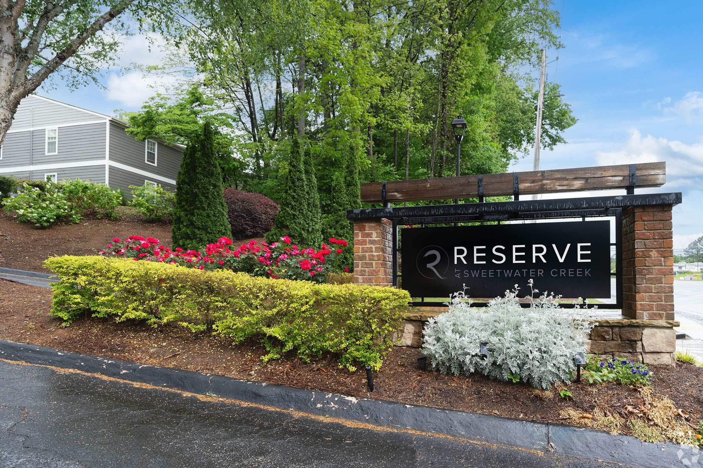 A sign for "Reserve at Sweetwater Creek" surrounded by colorful flower beds and shrubs. The scene features a clear blue sky and well-maintained greenery, indicating a welcoming residential community.